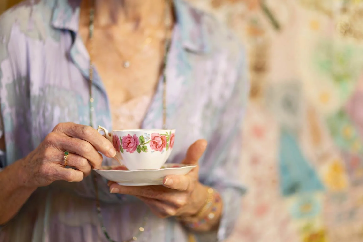 Picture of a woman' torso drinking in a dainty pink and white tea cup. She has a blue dress on and a faded tapestry in the background