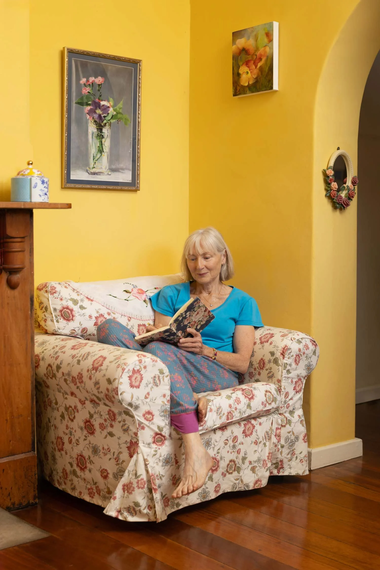 Penelope Lane, clinical psychologist, mindfulness teacher & ageing coach, sitting on a floral sofa, reading a book in a yellow-walled living room.
