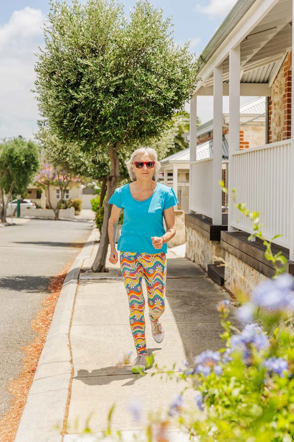 Penelope Lane, healthy ageing & lifestyle coach, jogging on a sidewalk in a sunny neighborhood, wearing red sunglasses, a blue top, and colorful floral leggings.