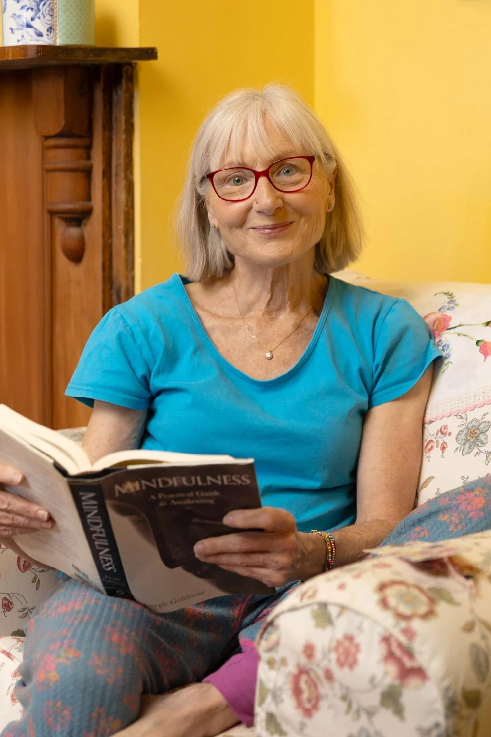 Penelope Lane, healthy ageing & lifestyle coach, wearing glasses and a blue shirt sitting on a floral-patterned sofa, holding a book. She is smiling at the camera in a cozy, colorful room with yellow walls and wooden furnishings.