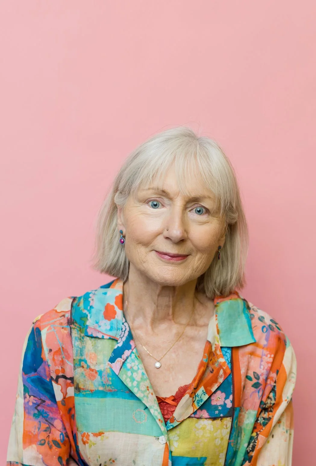 Penelope Lane, clinical psychologist & healthy ageing coach, wearing colorful earrings and a vibrant patchwork shirt, against a pink background.
