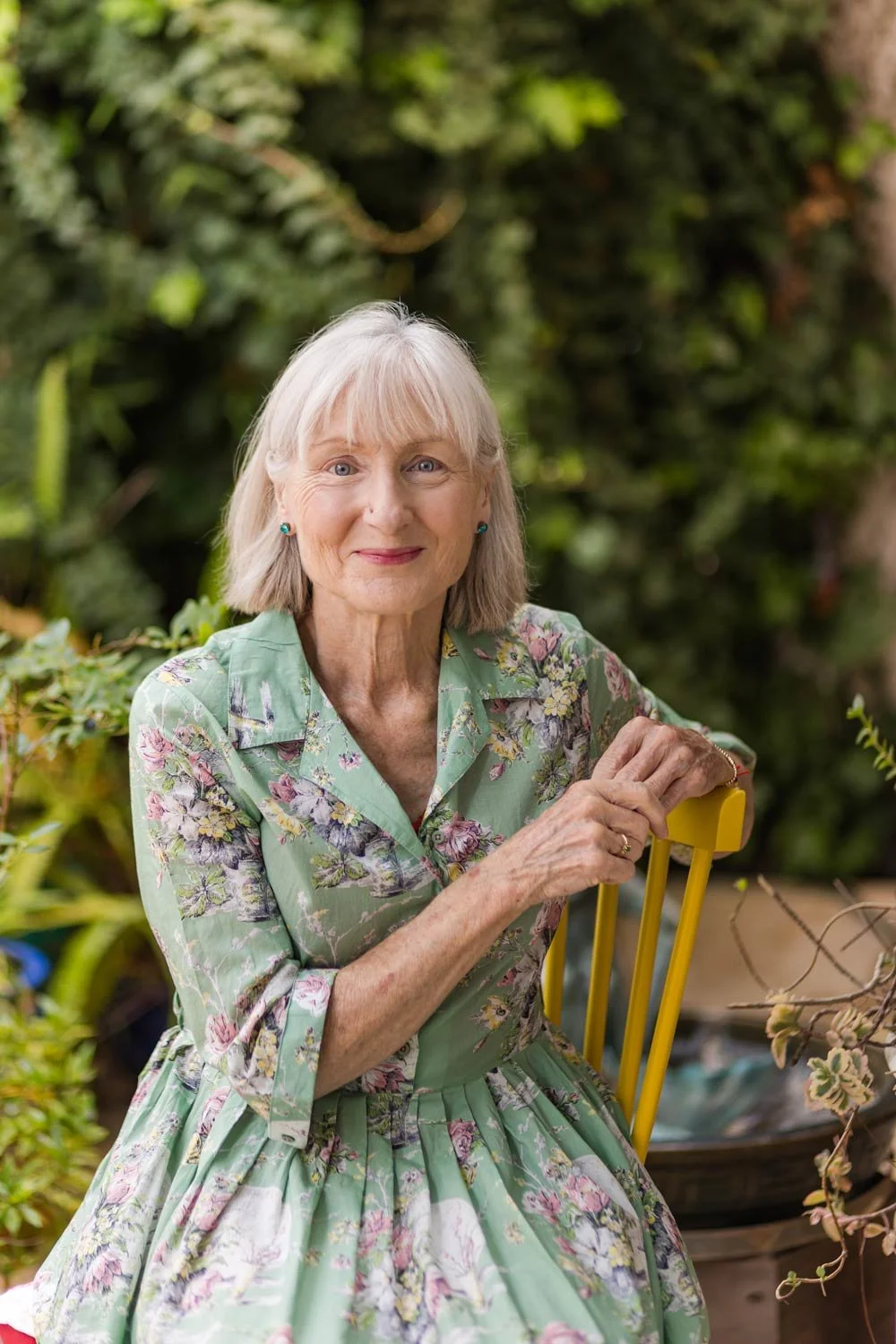 Penelope Lane, healthy ageing & lifestyle coach, wearing a floral green dress, sitting outdoors on a yellow chair and greenery in the background.