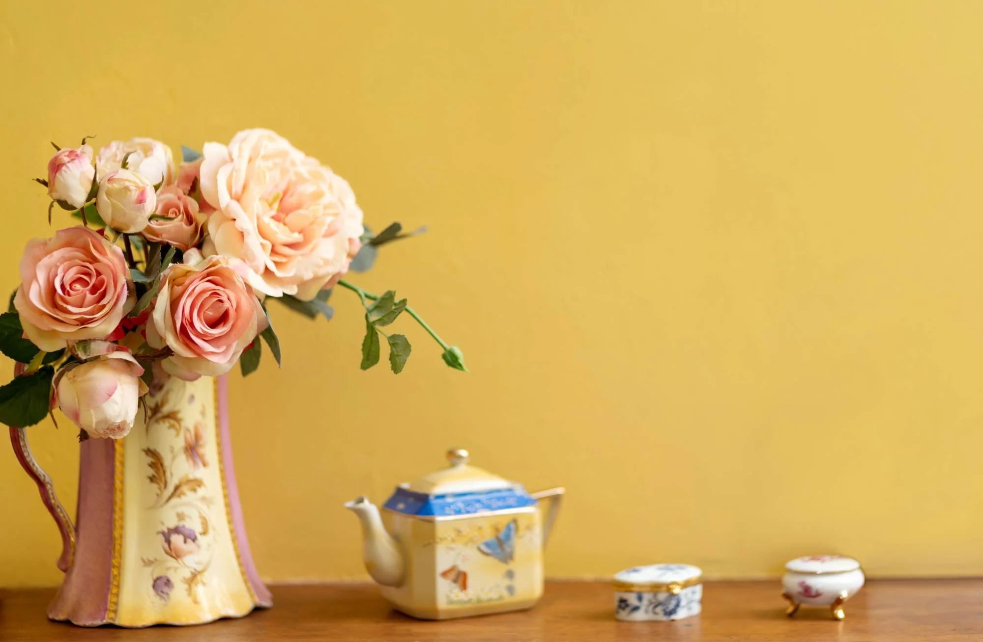 A floral arrangement of pink and cream roses in a decorative vase next to a teapot and small decorative boxes on a wooden surface against a yellow wall.