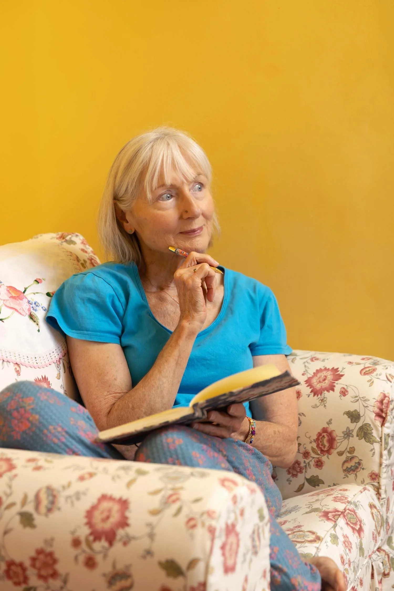 Penelope Lane, clinical psychologist, mindfulness teacher & ageing coach, sitting on a floral sofa with a yellow wall background, holding a notebook and a pen, looking thoughtful.
