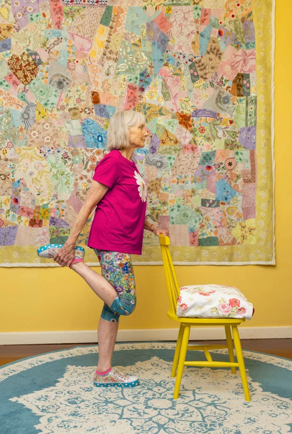 Penelope Lane performs a stretch while standing on one leg, holding her foot behind her back with one hand and using a yellow chair for support with her other hand. She stands in front of a colorful patchwork quilt hanging on a yellow wall.
