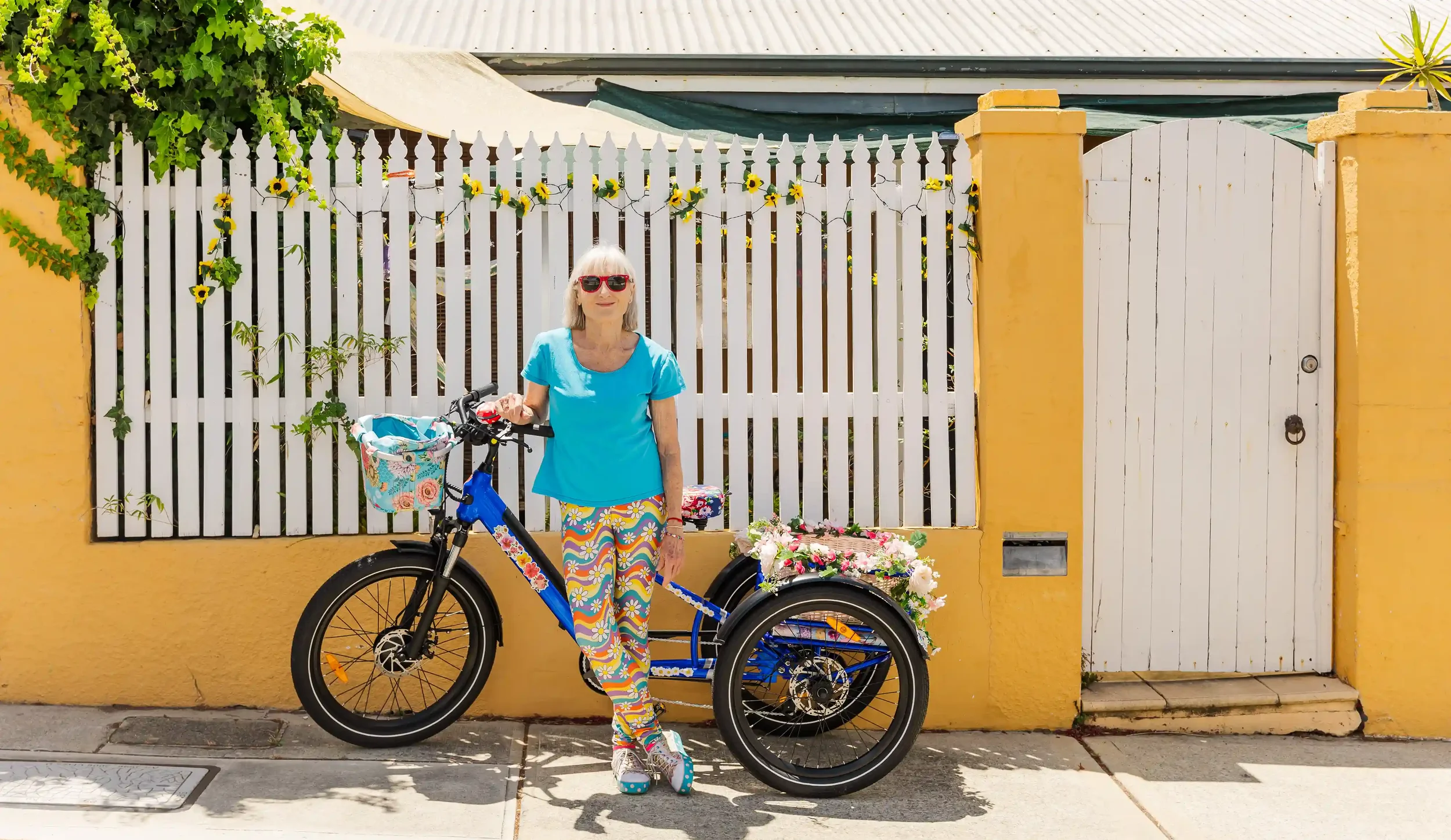 Penelope Lane wearing sunglasses, a blue shirt, and colorful patterned pants standing next to a blue bicycle with a floral basket and a rear cargo box filled with flowers, in front of a yellow wall with a white fence and gate.