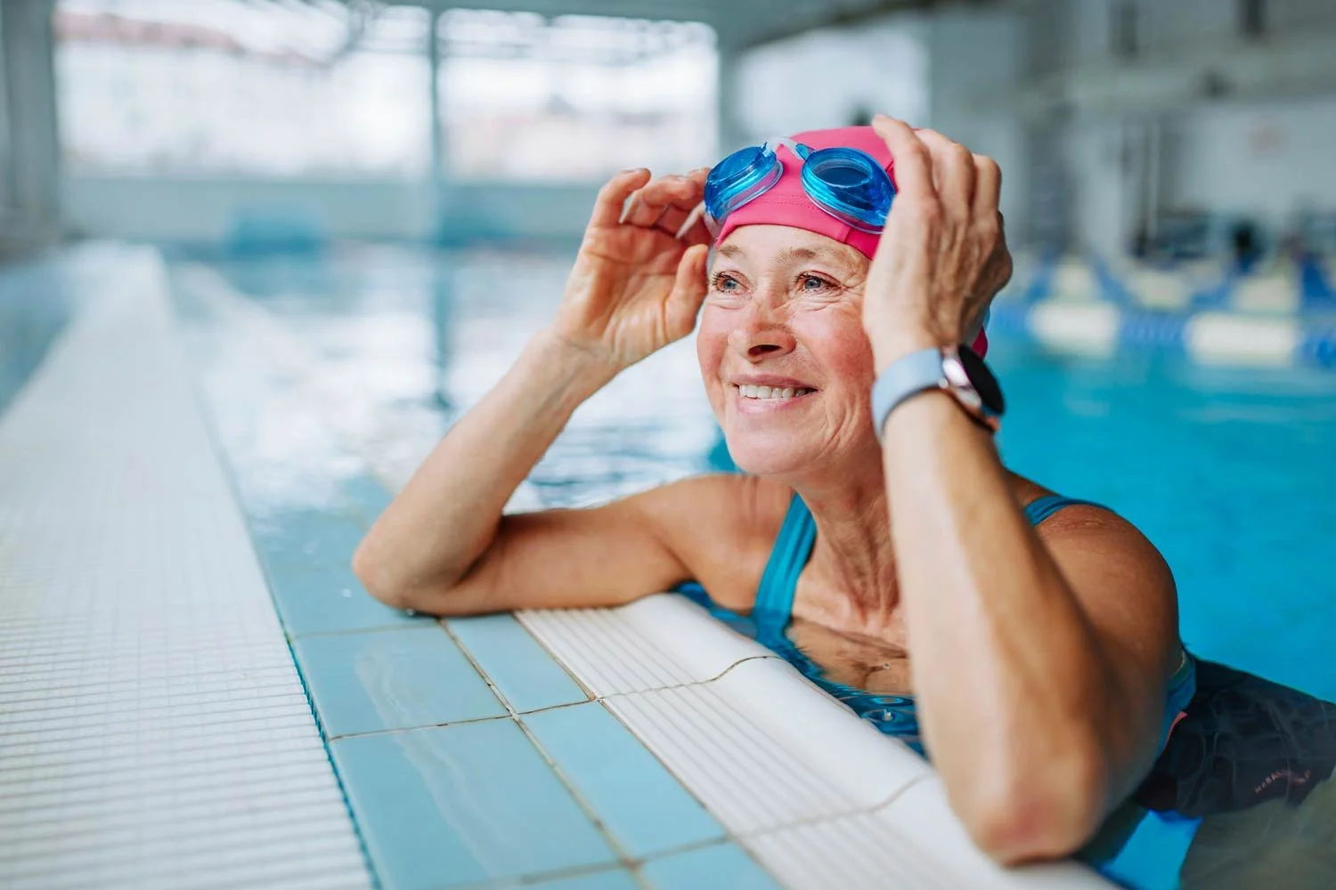 Elderly woman smiling and leaning on the edge of a pool with a pink swimmers cap and blue goggles.