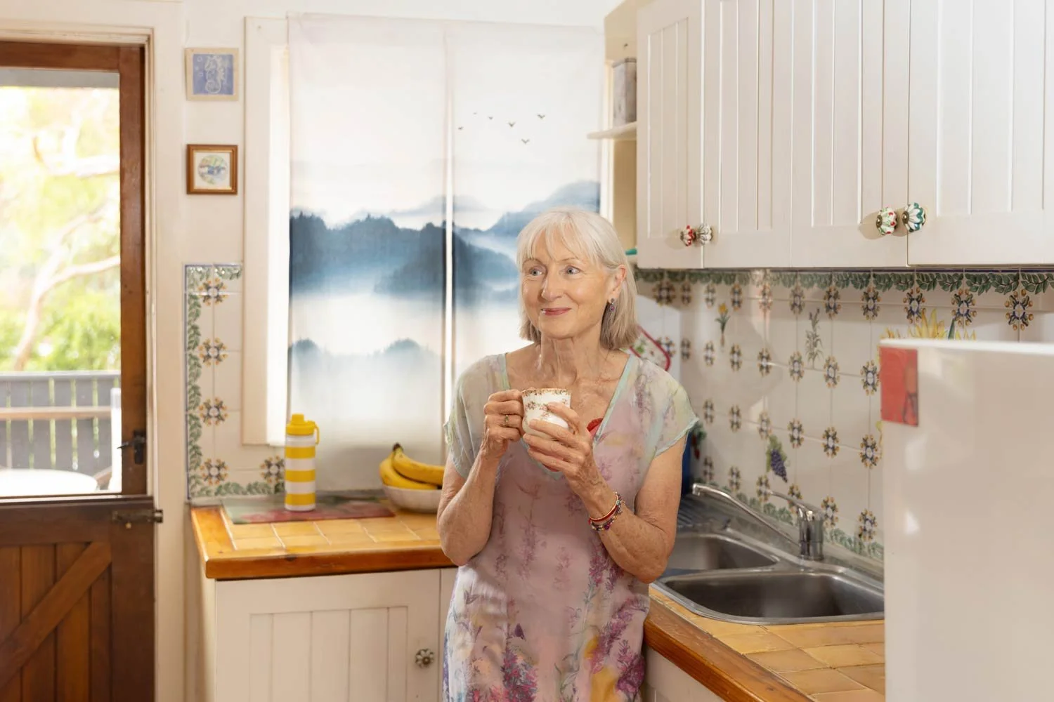 Woman in her kitchen with a cup of tea smiling at someone cheekily in the distance. Sunlight coming through the back door.