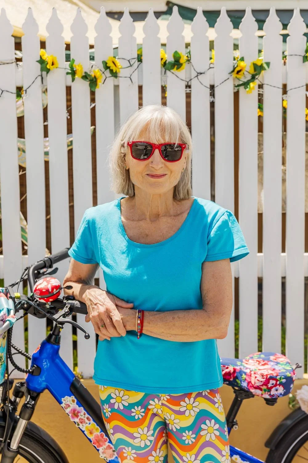 Penelope Lane, healthy ageing coach, wearing sunglasses, a blue t-shirt, and colorful floral-patterned pants, standing next to a bicycle with a floral saddle bag in front of a white picket fence decorated with yellow sunflower string lights.