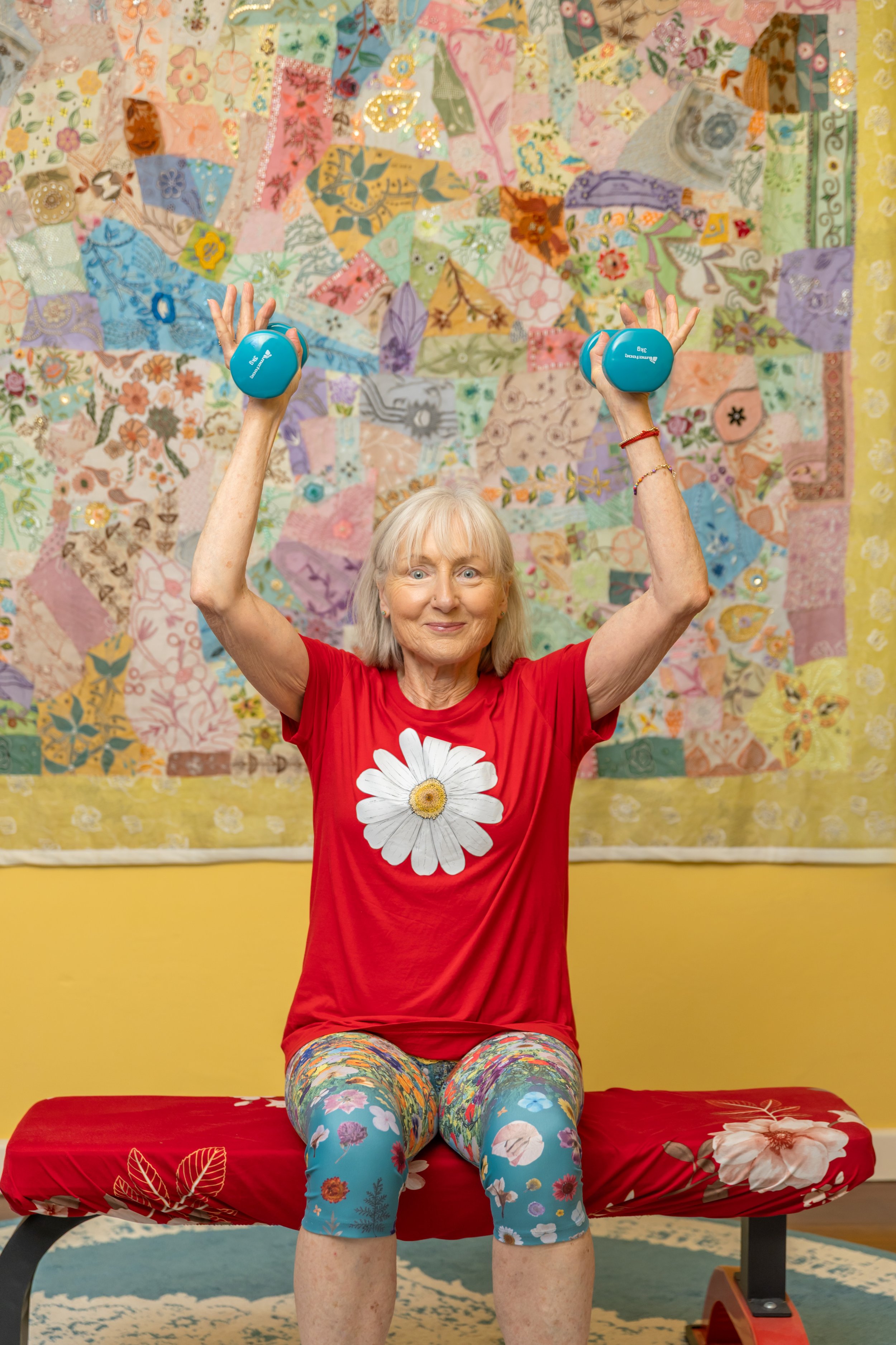 Woman on exercise bench indoors performing shoulder presses with blue dumbells. She's smiling and enjoying the exercise.