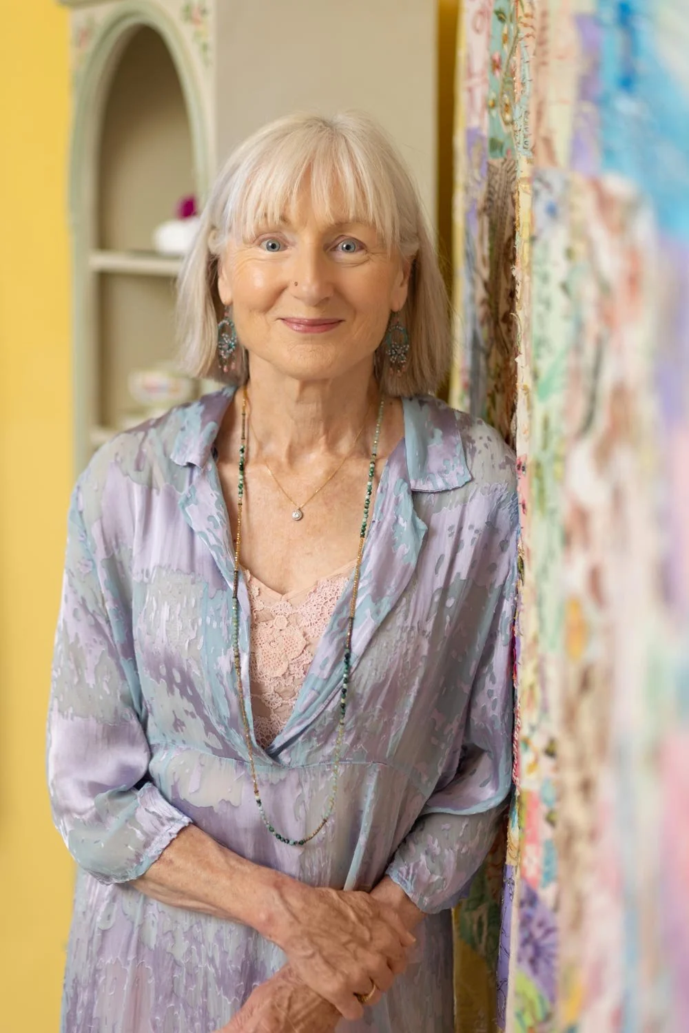 Older women standing smiling with grey hair leaning against a tapestry.