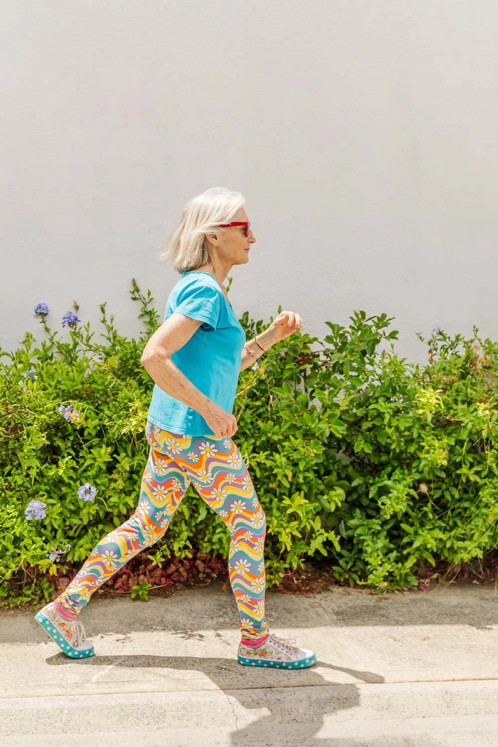Penelope Lane, healthy ageing & lifestyle coach, jogging outdoors on a sunny day, wearing sunglasses, a turquoise t-shirt, multicolored floral patterned leggings, and colorful sneakers in front of green bushes and a plain wall.