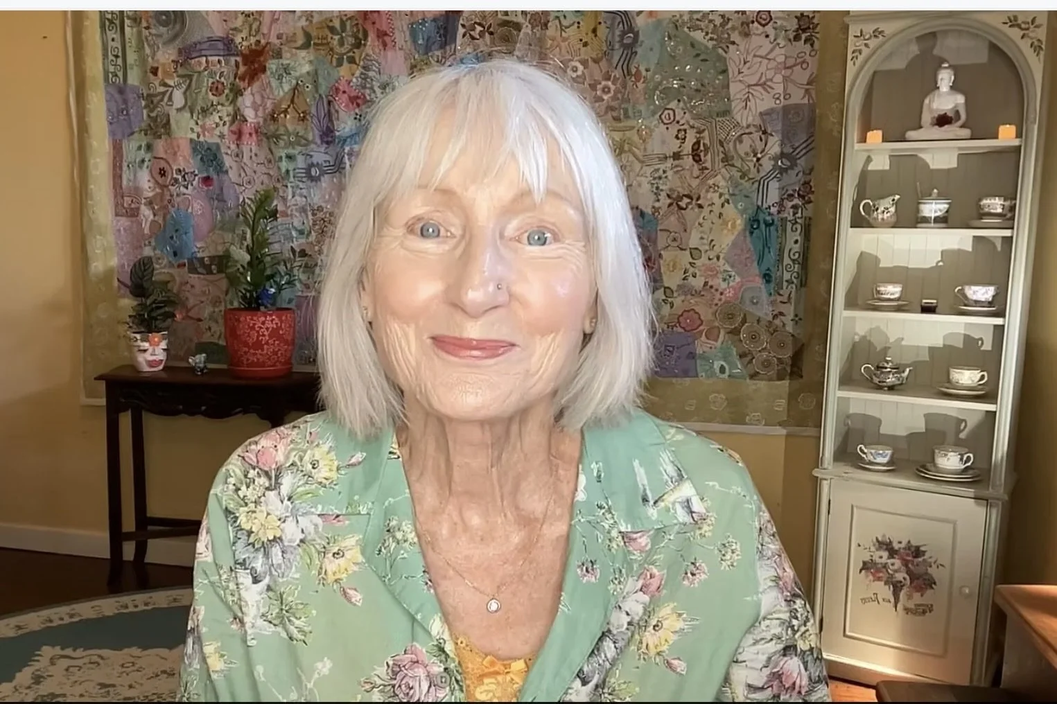 Older woman smiling sitting in her loungeroom with a green floral dress on with a yellow camisole.
