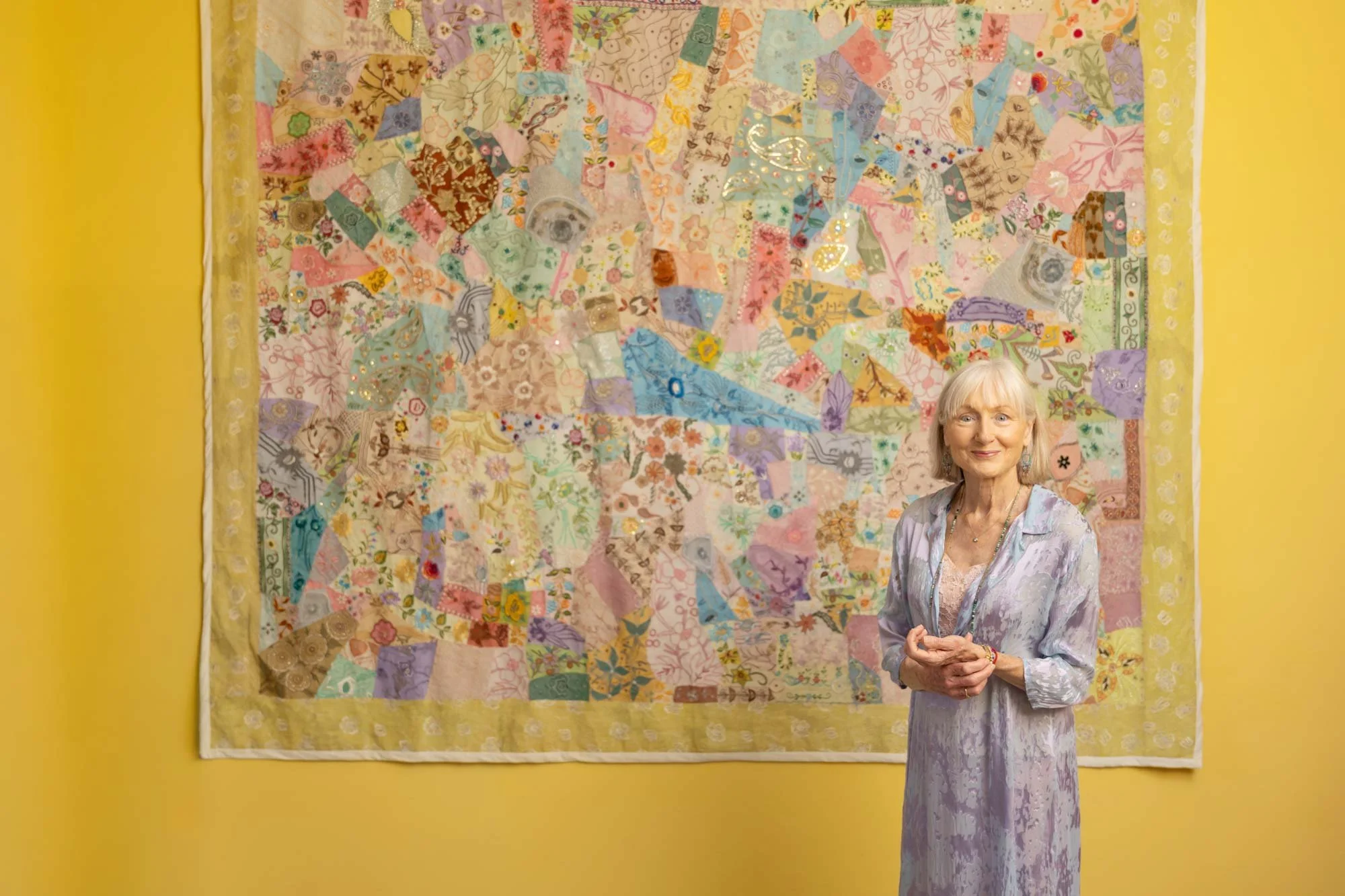 Penelope Lane, healthy ageing & lifestyle coach, standing in front of a colorful patchwork quilt hanging on a yellow wall. She is smiling and wearing a pastel dress with a silky, patterned fabric.