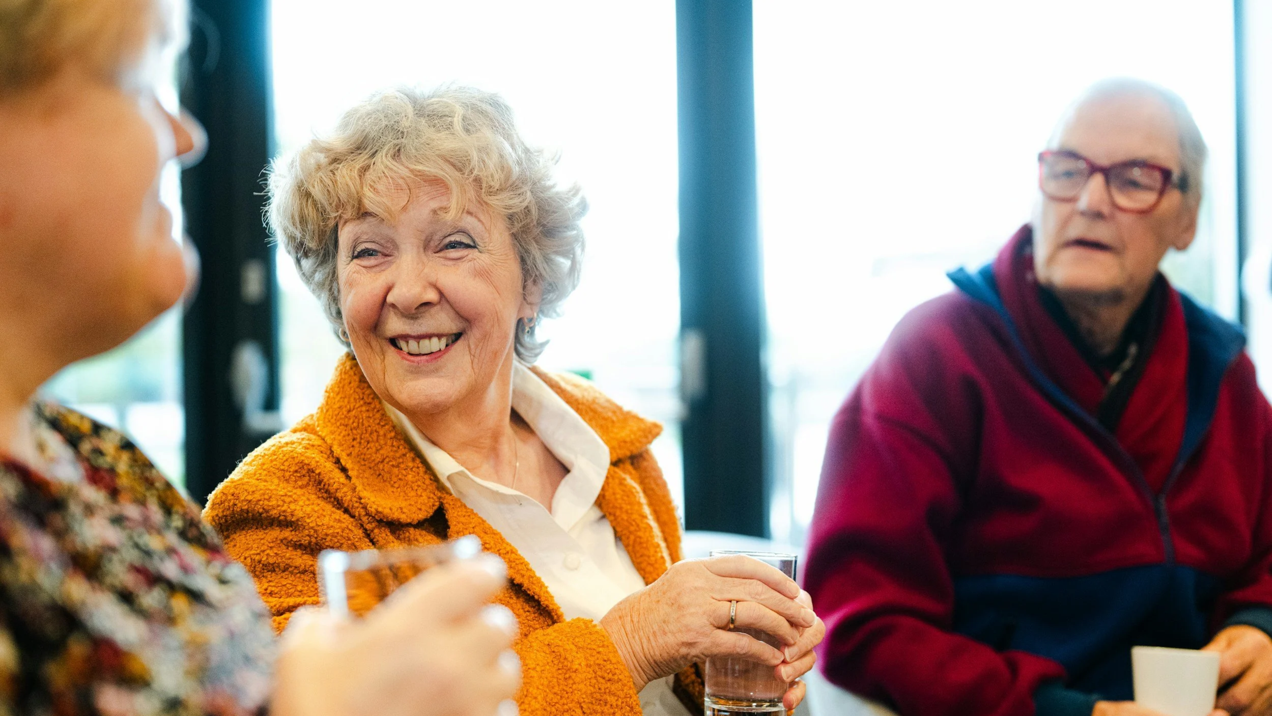 Women with grey hair, orange jumper sharing time with other women holding a glass of water