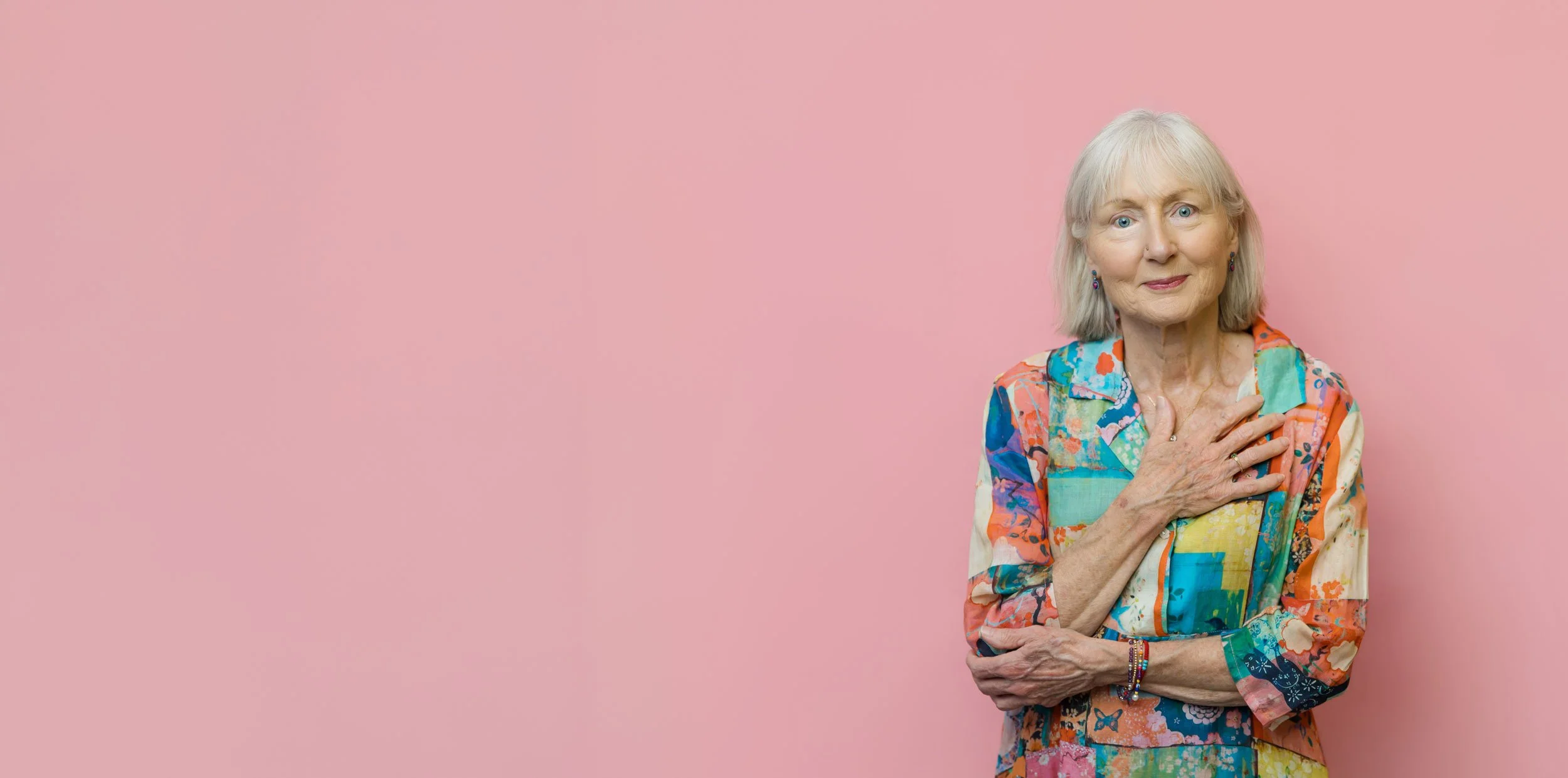 Penelope Lane, clinical psychologist & healthy ageing coach, wearing colourful, patterned clothing, standing against a pink background, with her right hand over her heart.