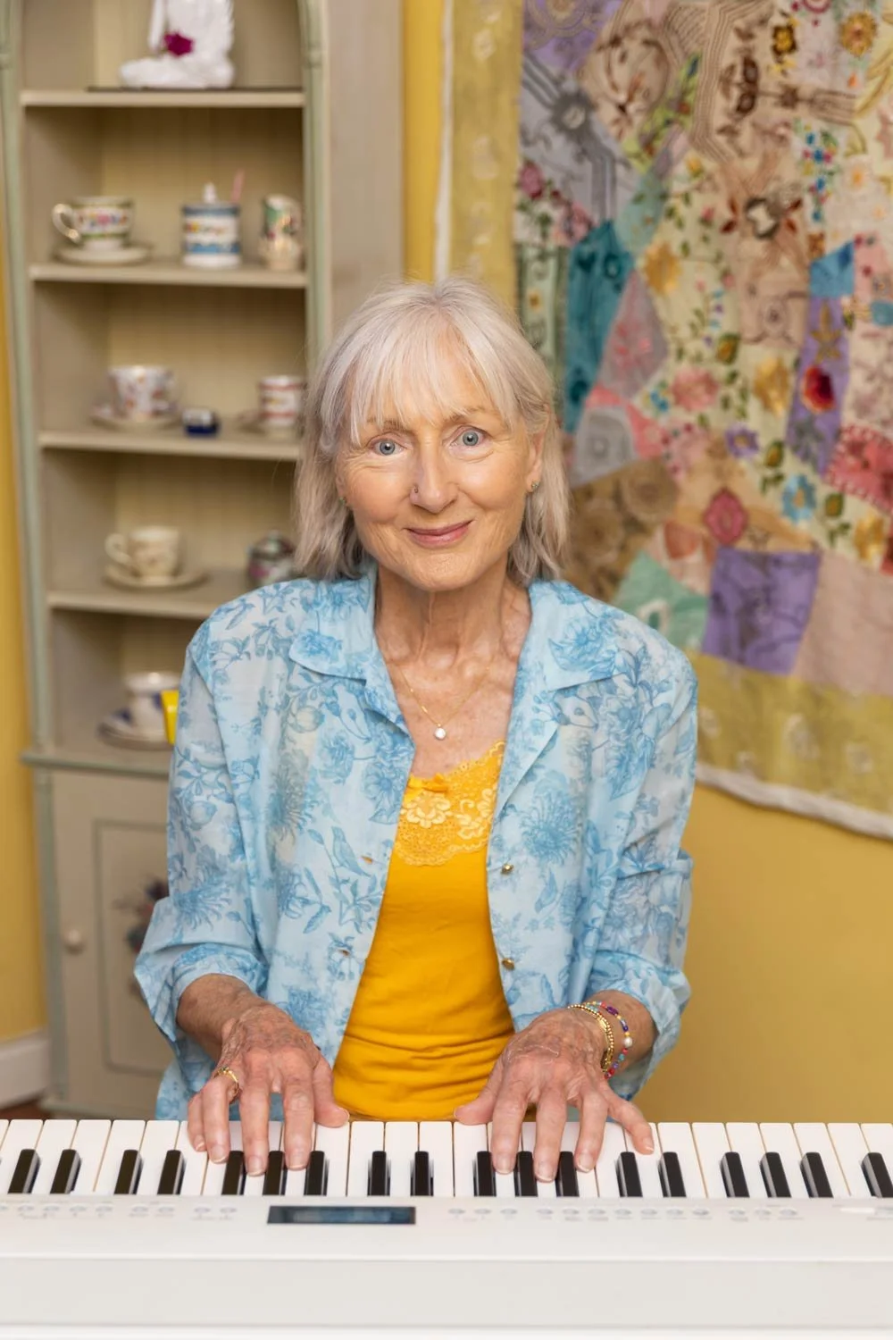 Penelope Lane, clinical psychologist, mindfulness teacher & ageing coach, playing a keyboard in a room decorated with a colorful quilt on the wall and a cabinet filled with teacups and dishes in the background.