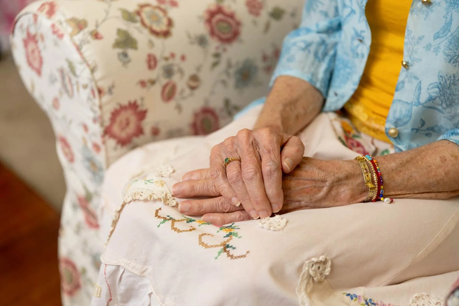 Close-up of Penelope Lane's hands resting on her lap while seated in a floral armchair.