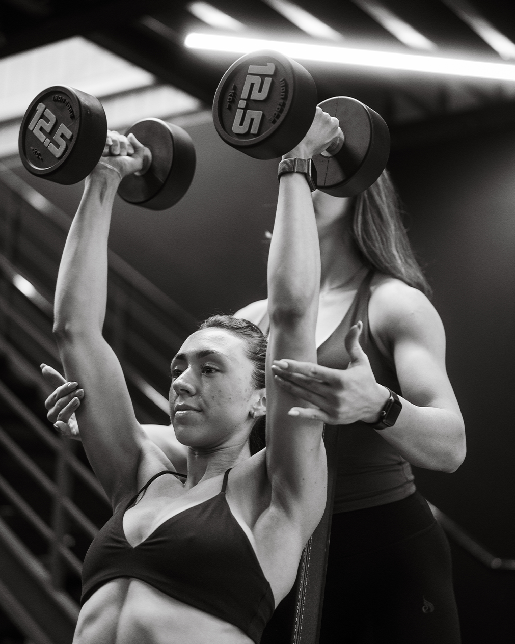 A woman doing a shoulder press exercise with dumbbells indoors, supervised by a trainer.