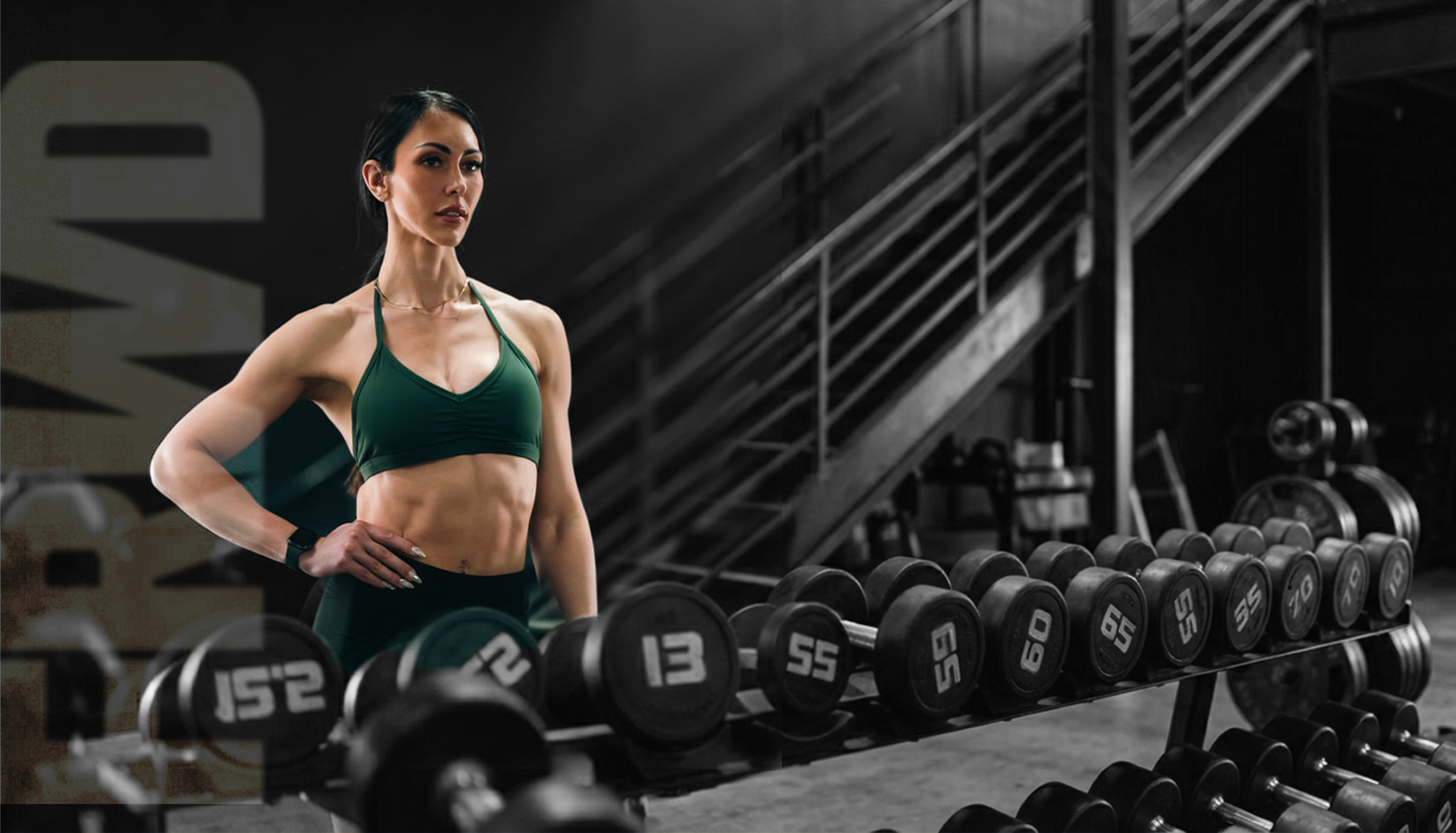 A fit woman in a green sports bra and black leggings stands with her hand on her hip in a gym, with rows of dumbbells in the foreground and a industrial staircase in the background.
