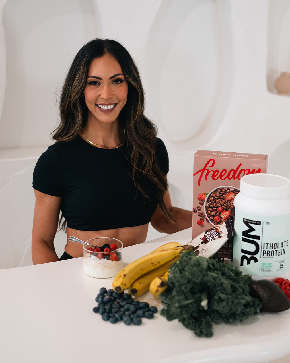 Smiling woman sitting at a table with healthy foods and supplements, including bananas, kale, blueberries, a berry yogurt, protein powder, and cereal.