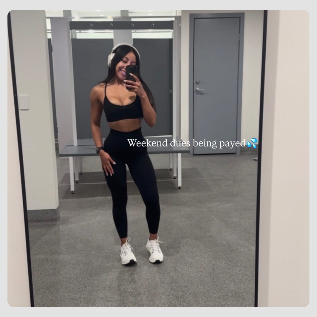 A woman in workout clothes taking a mirror selfie in a gym locker room.