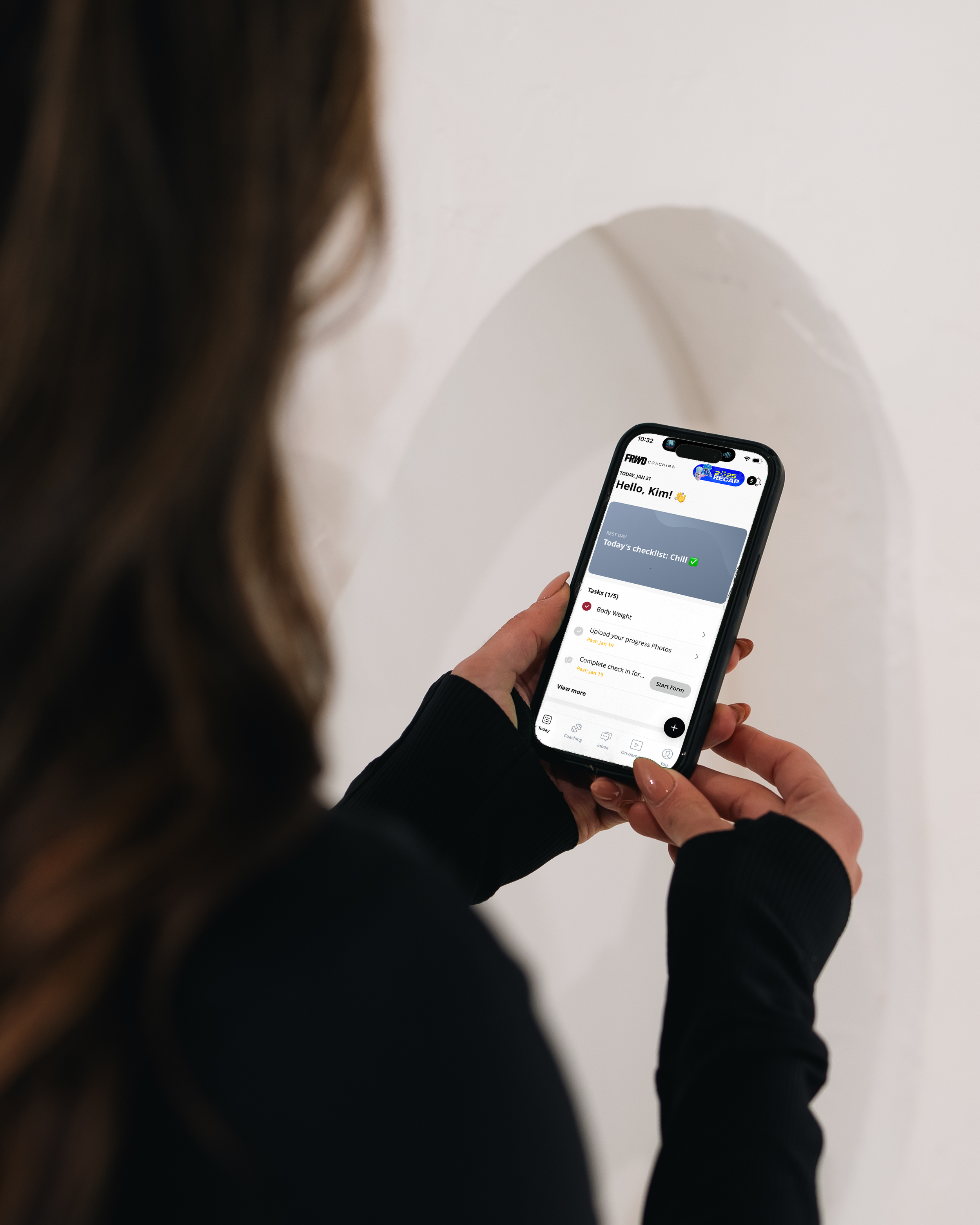 Person holding a smartphone displaying a fitness or coaching app with a checklist to be completed, including tasks like body weight, uploading progress photos, and checking in.