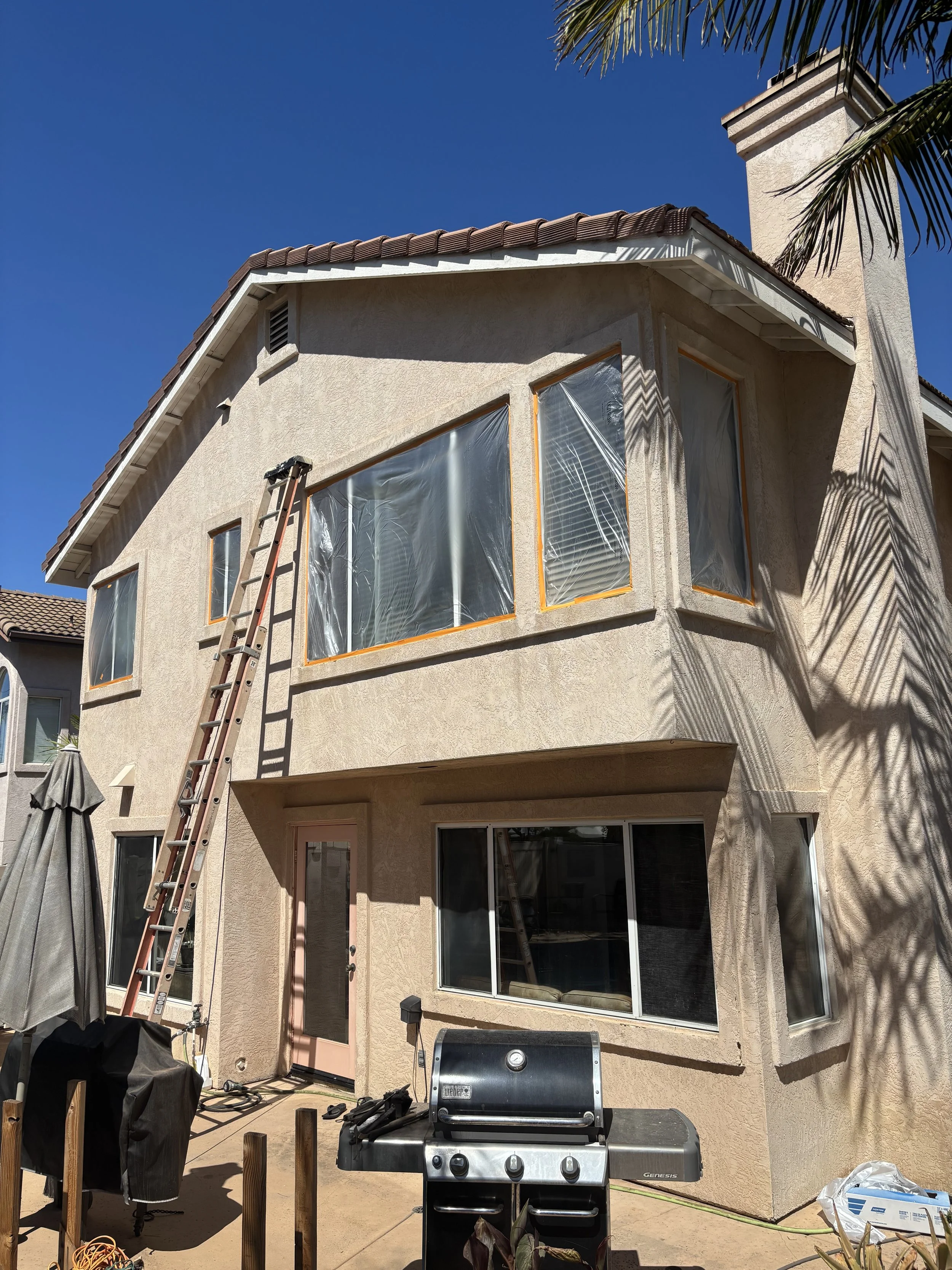 A two-story house under construction with plastic-covered windows, a ladder leaning against the wall, and a barbecue grill on the patio. Shadows of palm leaves are visible on the house wall.