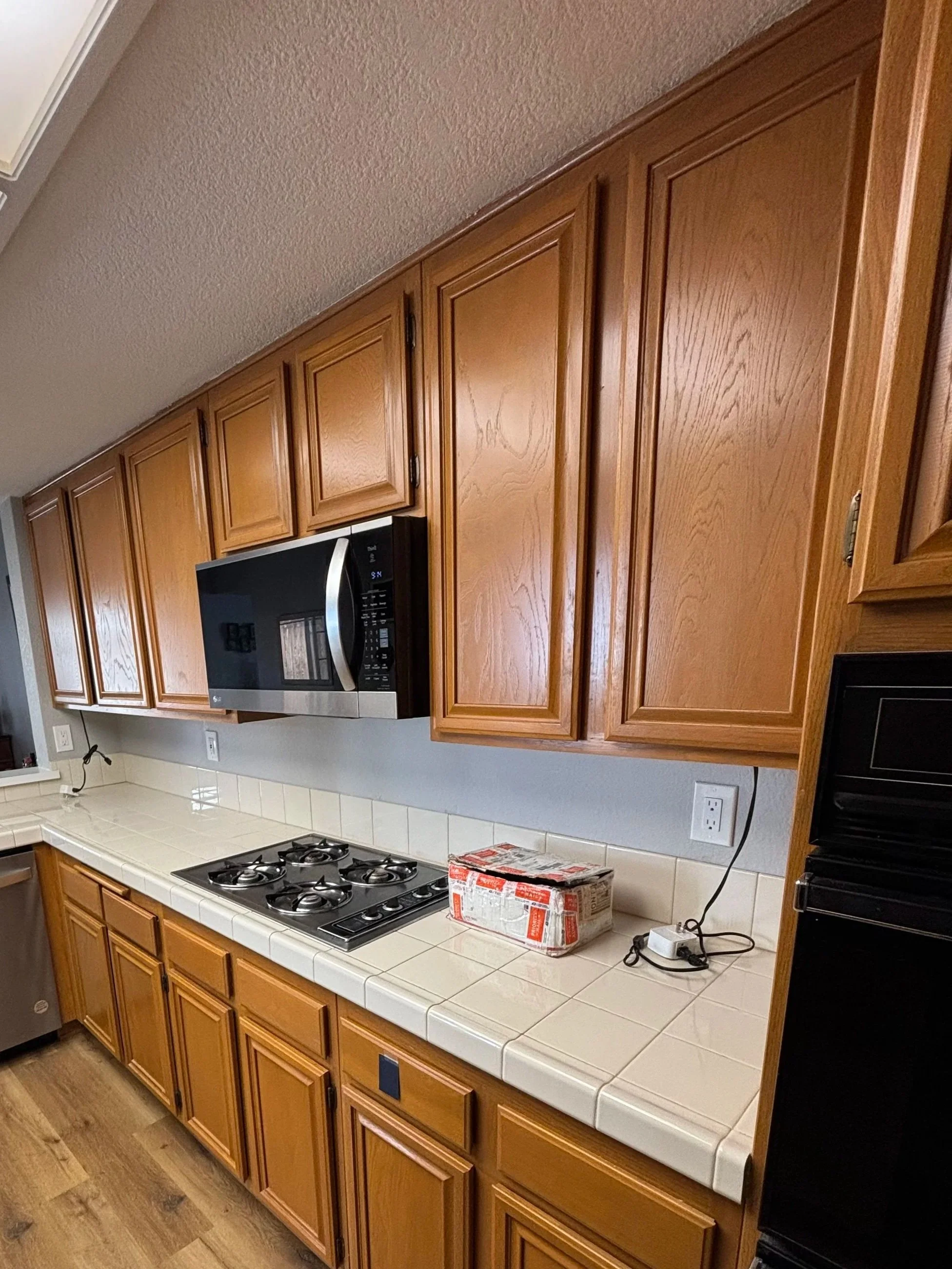 Kitchen with wooden cabinets, a microwave oven above the stove, a stove with four burners, a box of food or supplies on the counter, and electrical outlets, with a wooden floor.