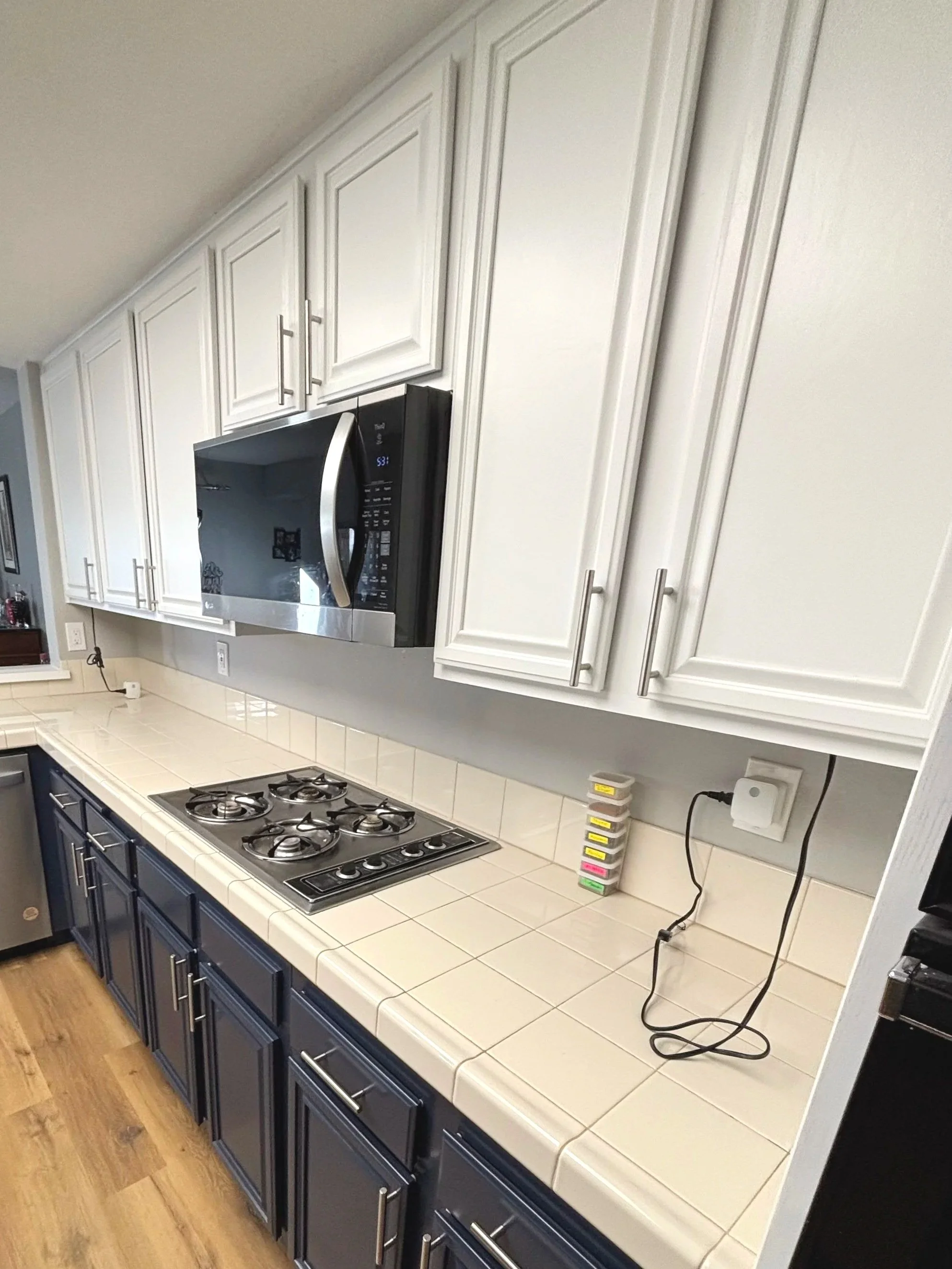 Kitchen countertop with white tiled surface, a black and stainless steel microwave mounted above, black cabinets below, a built-in gas stovetop, and a stack of labeled spice containers along with an electrical outlet with connected cords.