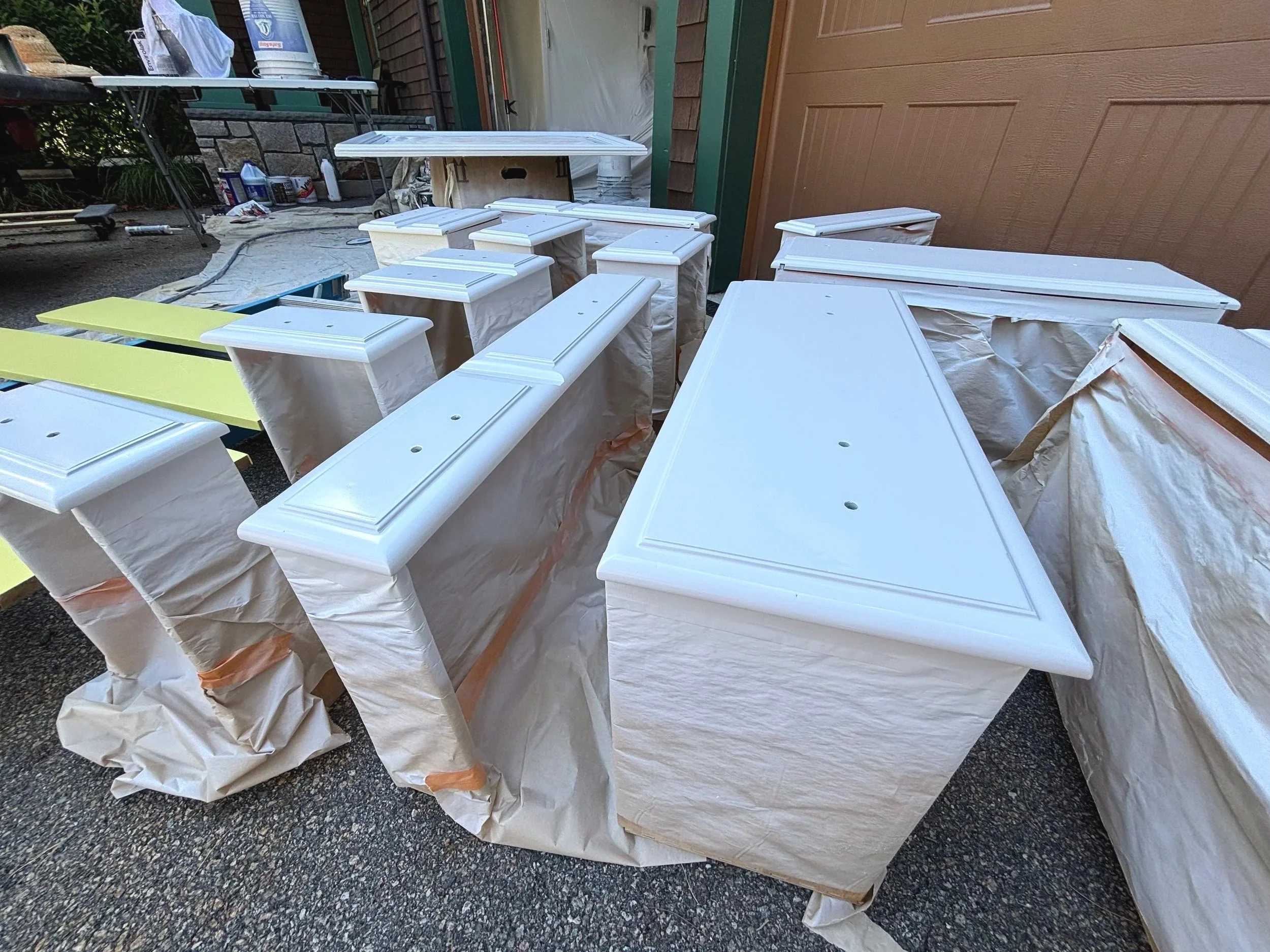 Multiple furniture pieces, including cabinets and tables, are painted white and are drying outdoors, covered with protective paper and tape.