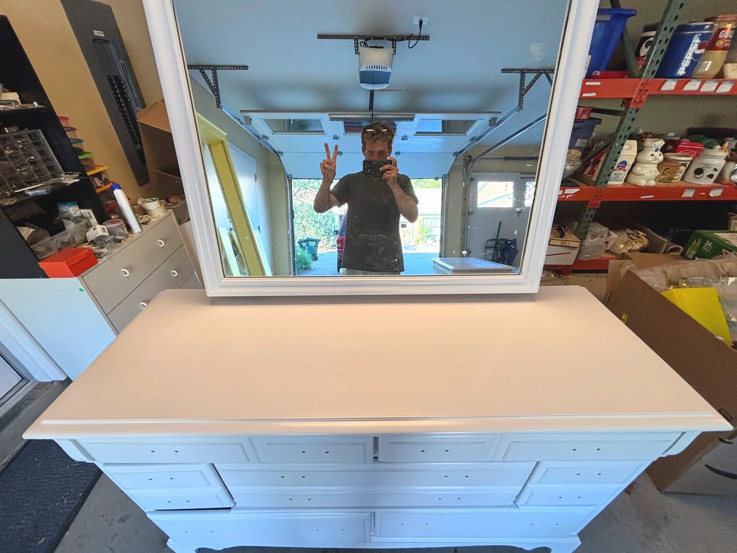 A person taking a selfie in a mirror shows a garage with work shelves filled with boxes, jars, and building materials. The person is making a peace sign with one hand.