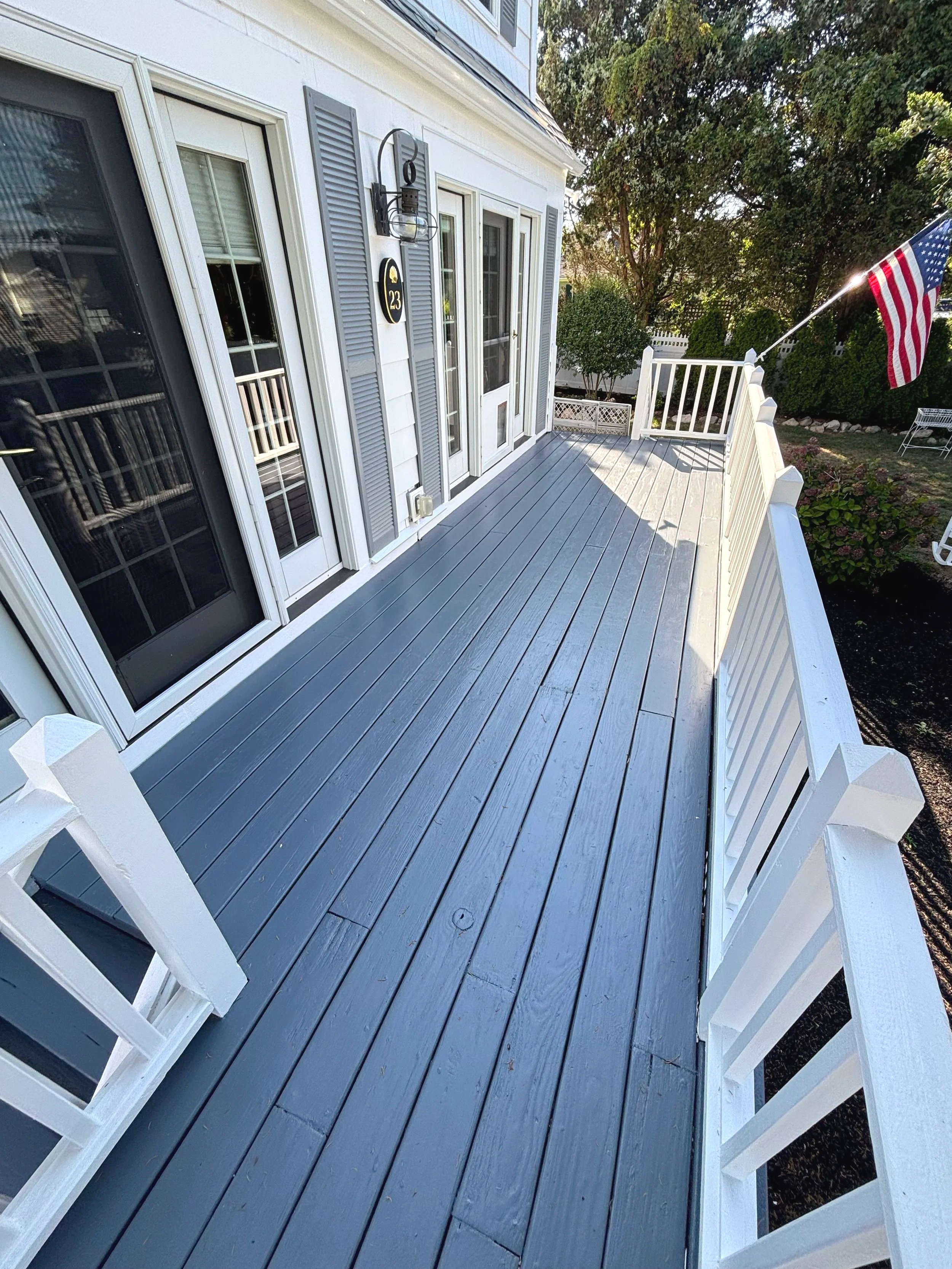 A view of a white house's back porch with gray wooden decking, white railing, and a door with sidelights, decorated with an American flag.