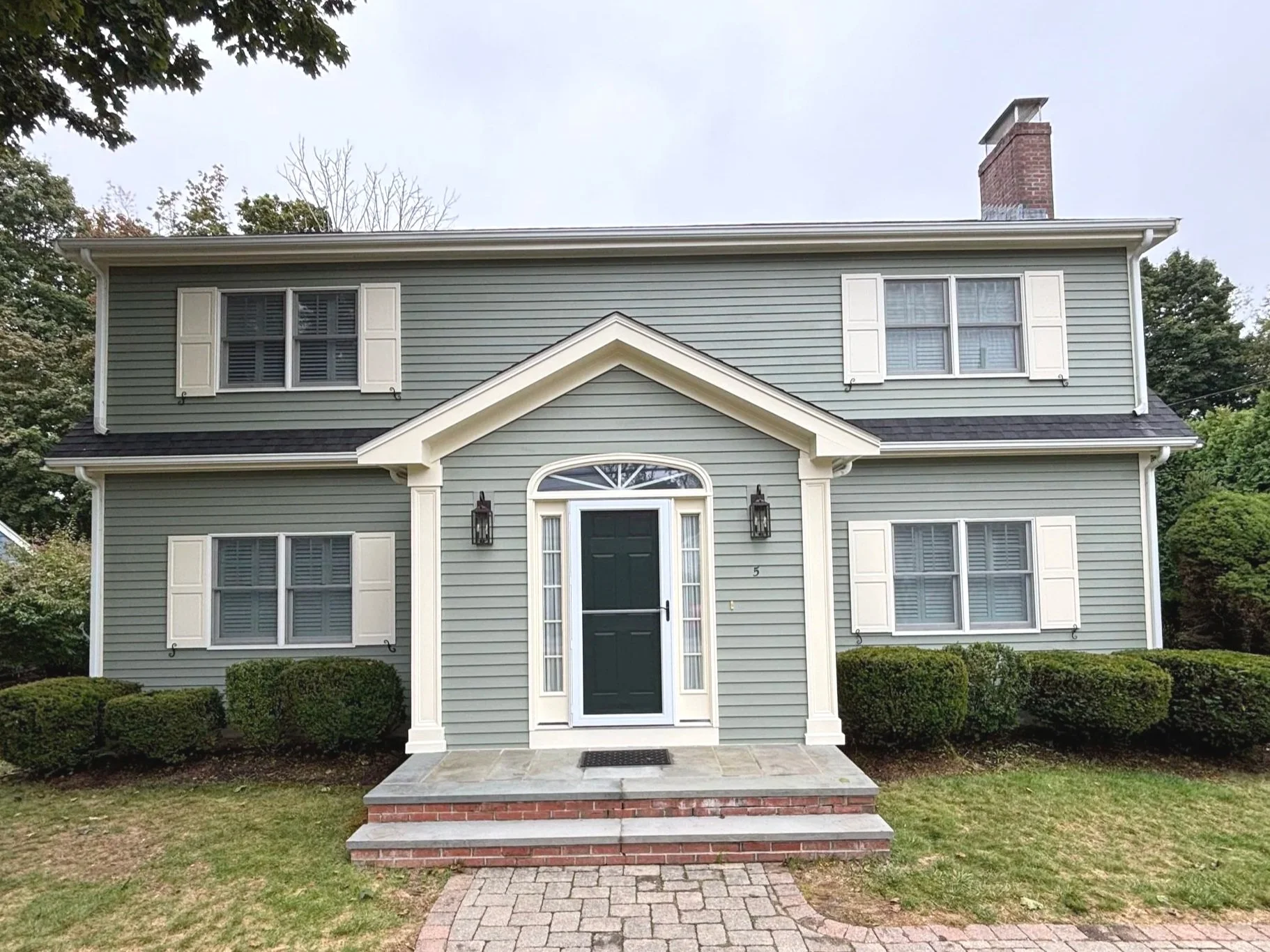 A two-story house painted in light gray with white window shutters and trim. The front door is dark green, with a semicircular window above and sidelights on each side. There are two black wall-mounted lanterns beside the door. The house has a small 