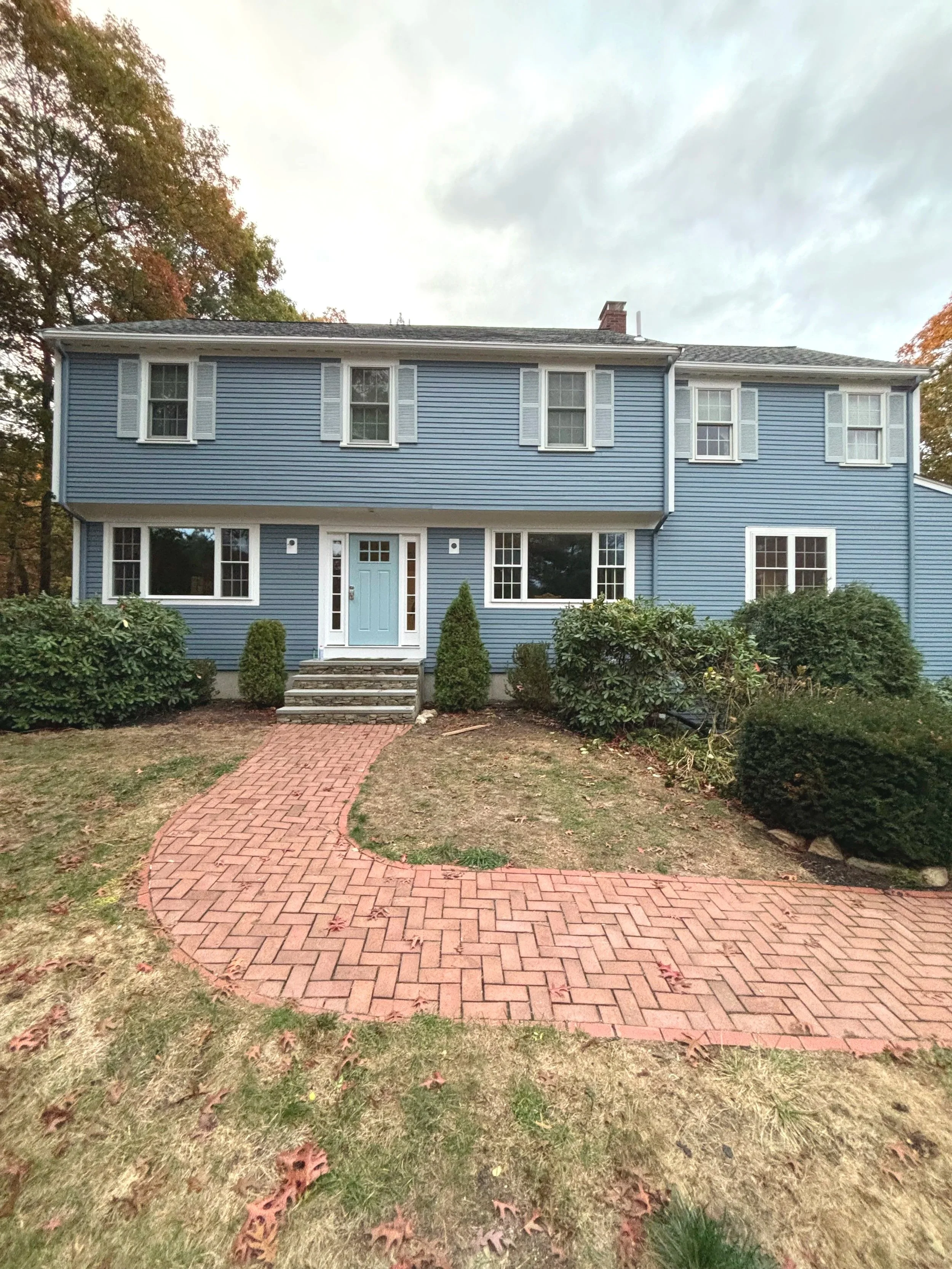 A blue two-story house with a brick walkway leading to the front door, surrounded by bushes and trees, under a cloudy sky.