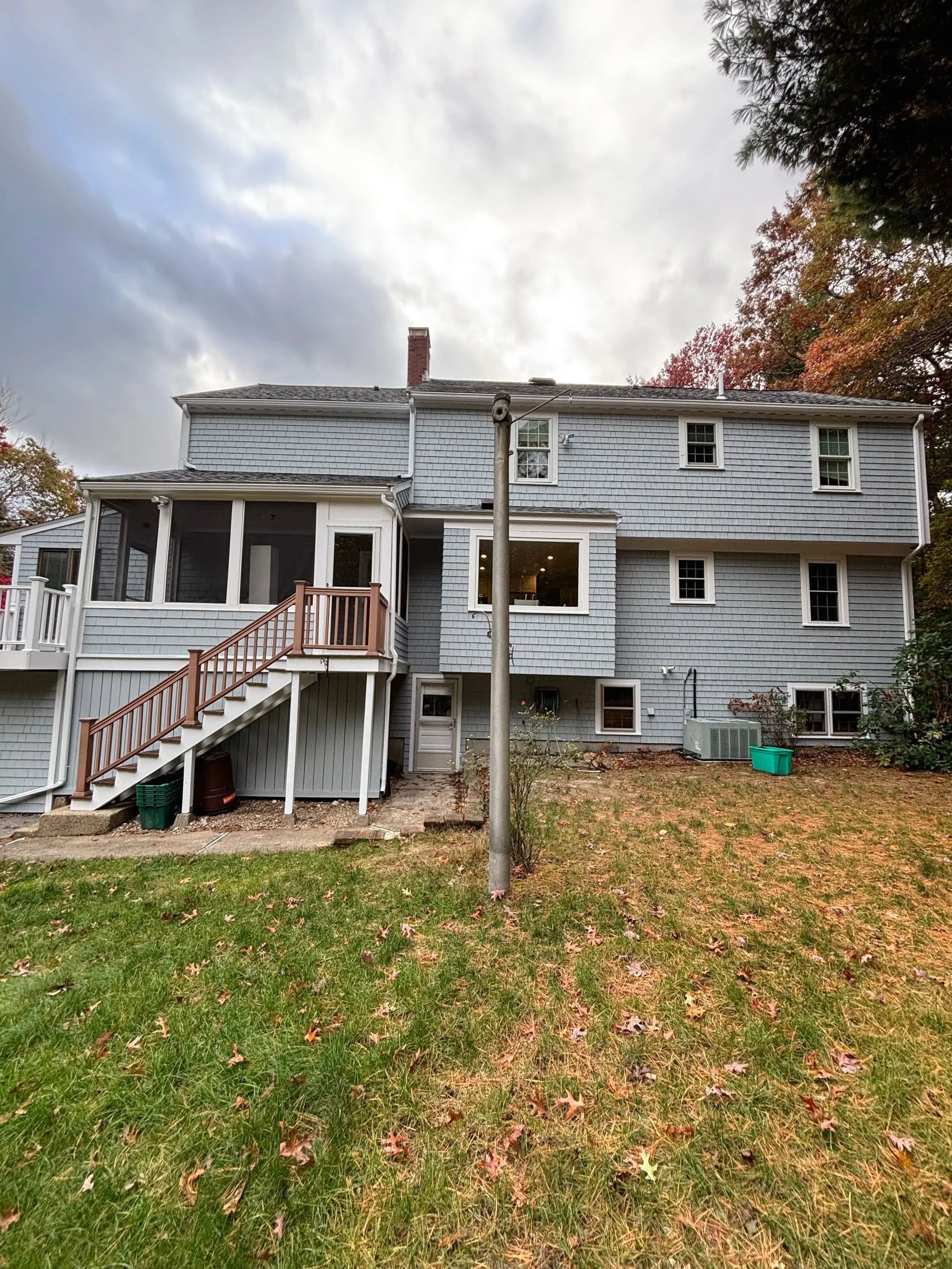 Backyard view of a multi-story house with gray siding, multiple windows, a small set of stairs leading to a porch, a utility pole in the foreground, and fall foliage on the trees.