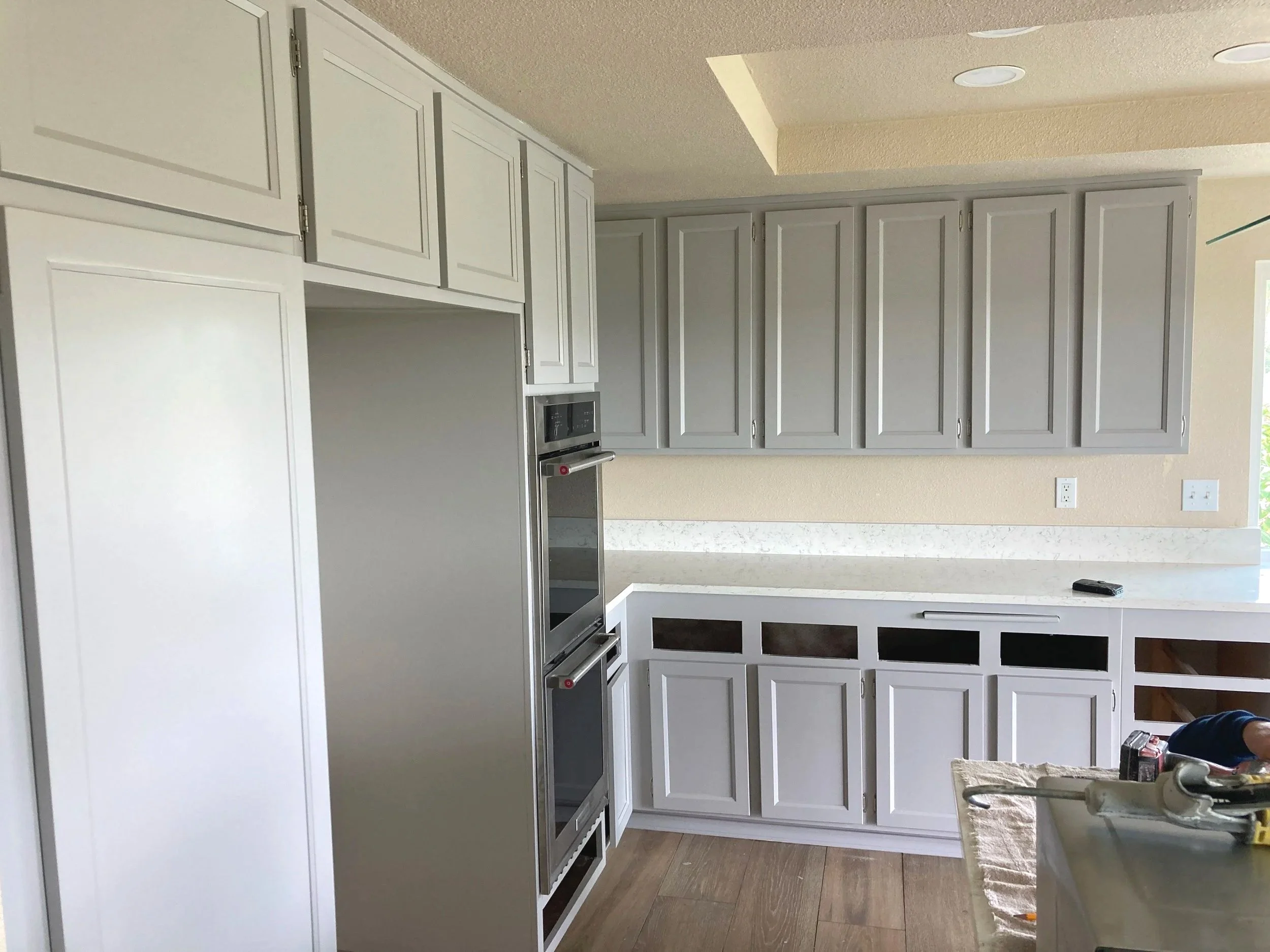 Kitchen with white cabinets, stainless steel double oven, and a white countertop. Some cabinet doors are open or missing, revealing dark space inside. There are electrical outlets on the wall and construction tools on the countertop.