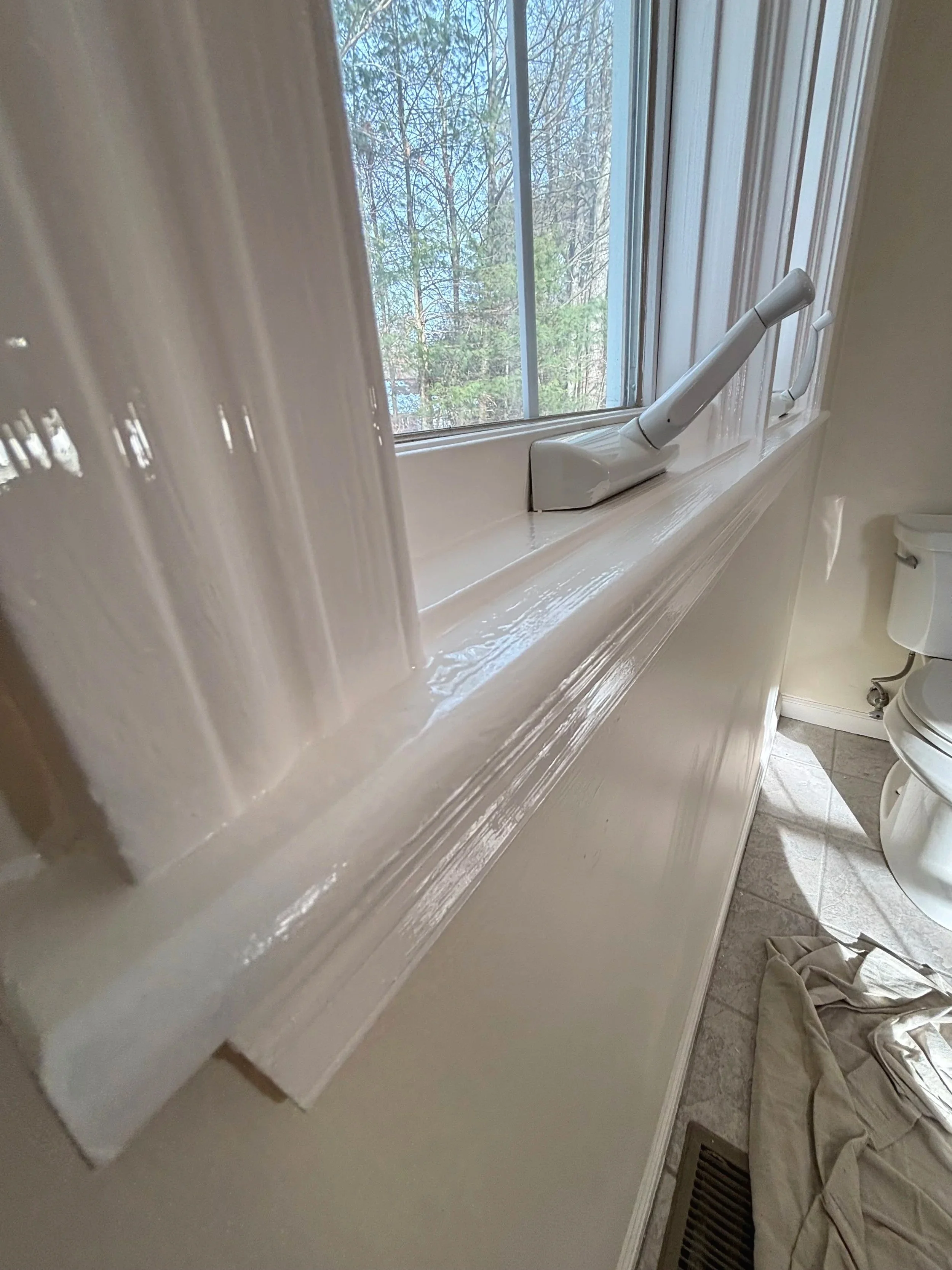 Close-up of a white window frame with a white window handle, overlooking trees outside in a bathroom with part of a toilet and a cloth on the floor.
