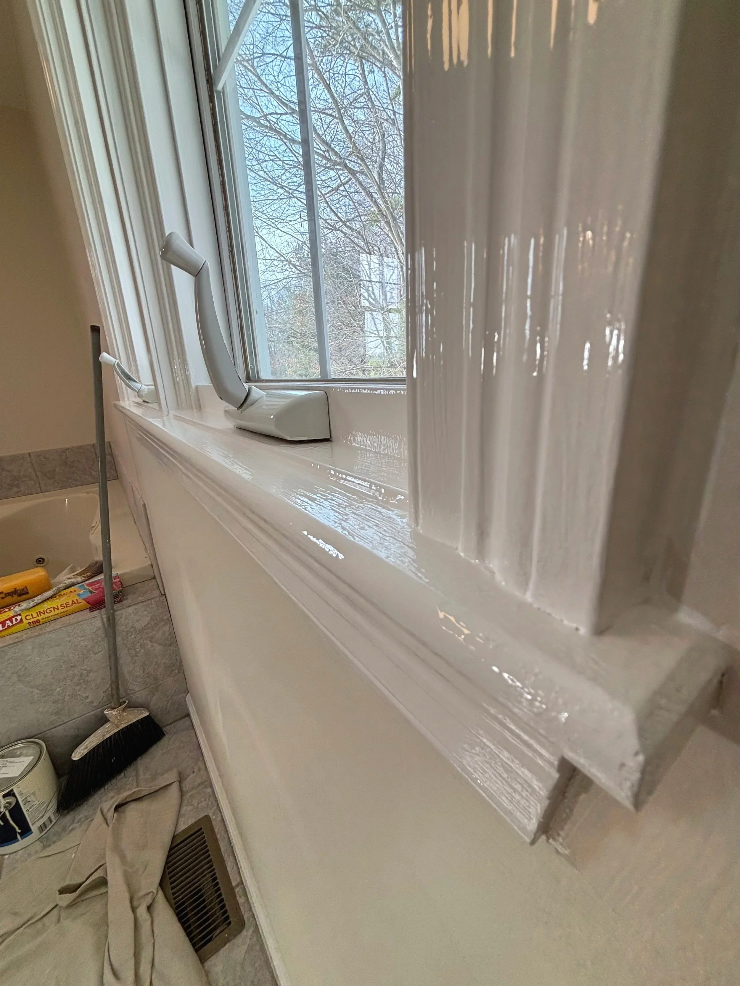 Close-up photo of a freshly painted white windowsill and trim in a room, with a window showing leafless trees outside in the background.