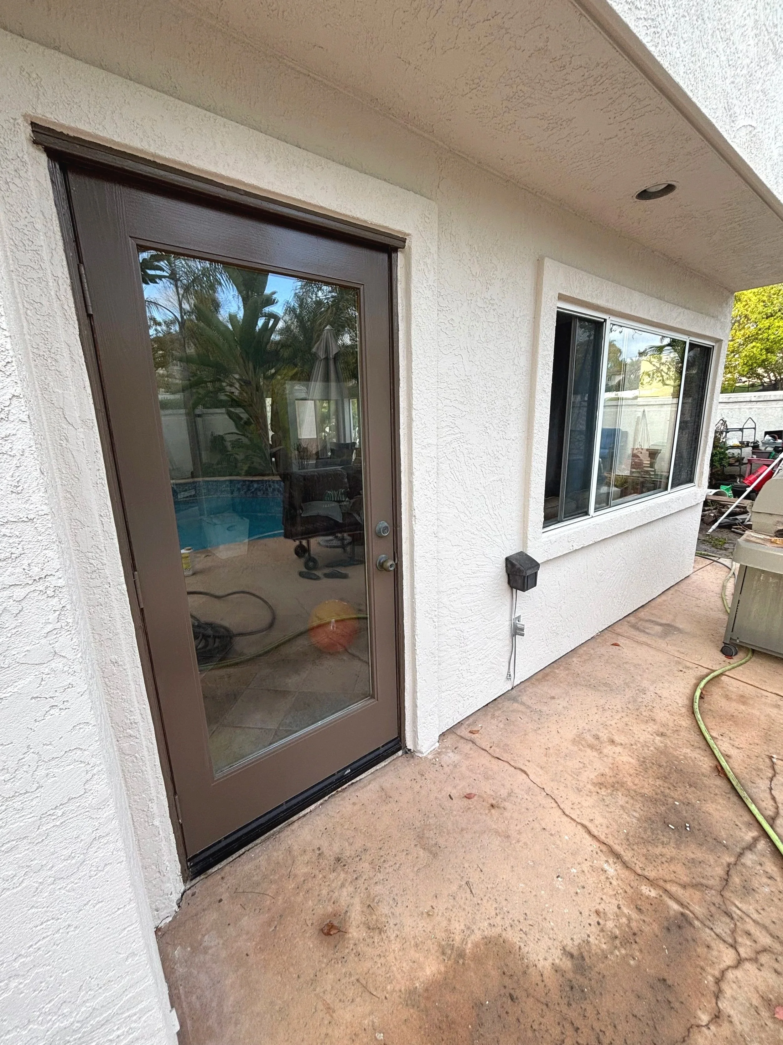Back patio area with a brown door with a glass window, white stucco walls, a large window, and typical outdoor items such as a garden hose, a tennis ball, and a portable barbecue grill.