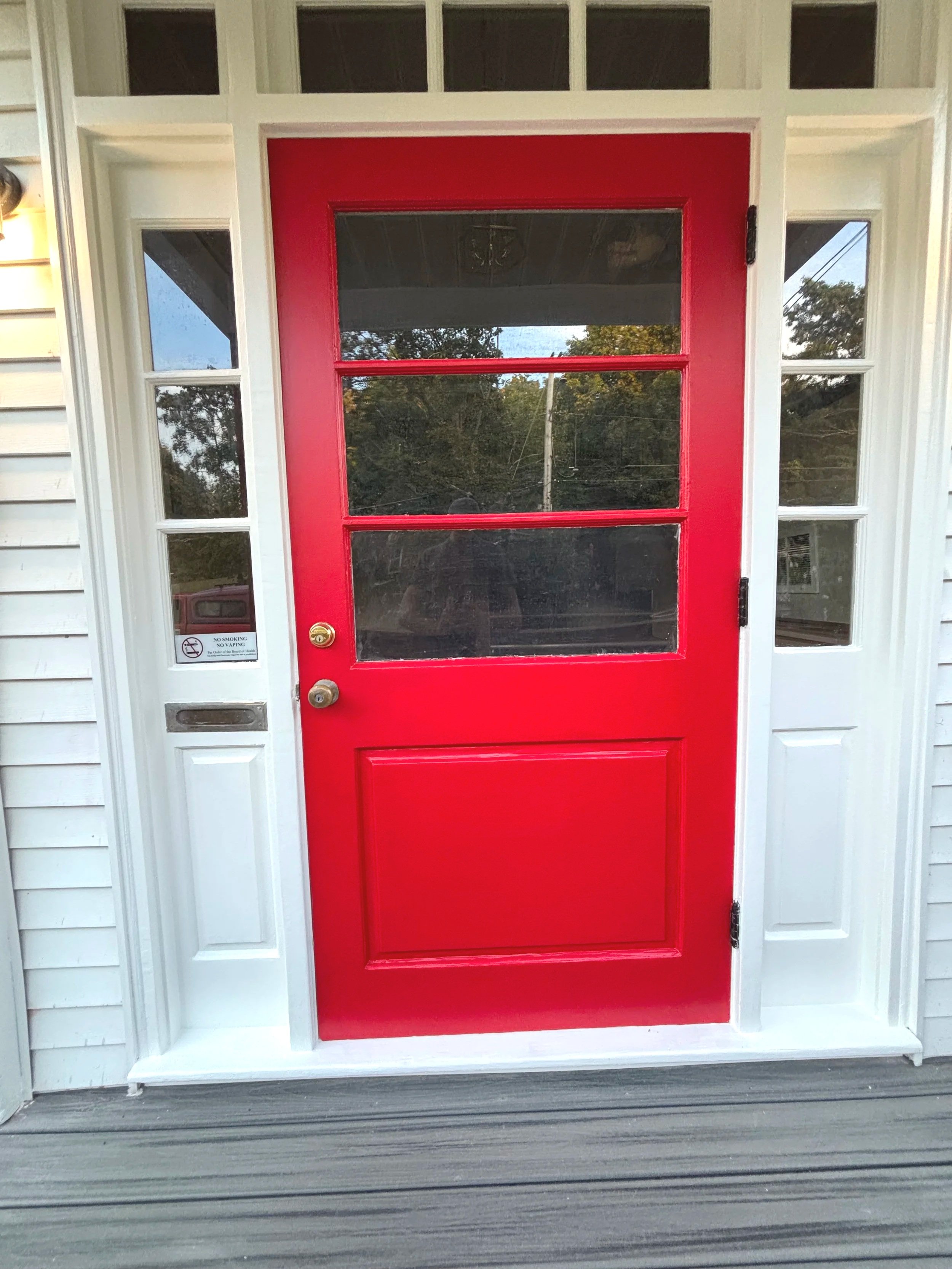 Red front door with three rectangular glass panels, surrounded by white trim and sidelights, with a gray wooden porch and siding.