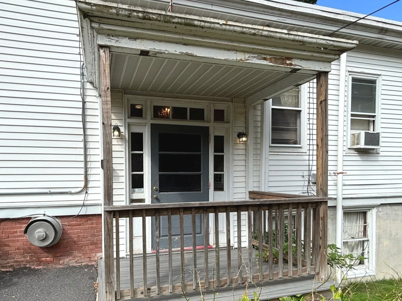 A small wooden porch with a railing and a door with a glass panel, attached to a white house with two windows, one with an air conditioning unit. The house's exterior has white siding and a brick foundation. There are two wall-mounted outdoor lights 