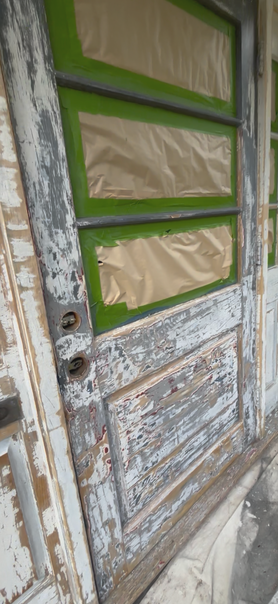 An old wooden door being renovated, with sections taped off with green painter's tape and brown paper for painting or staining.
