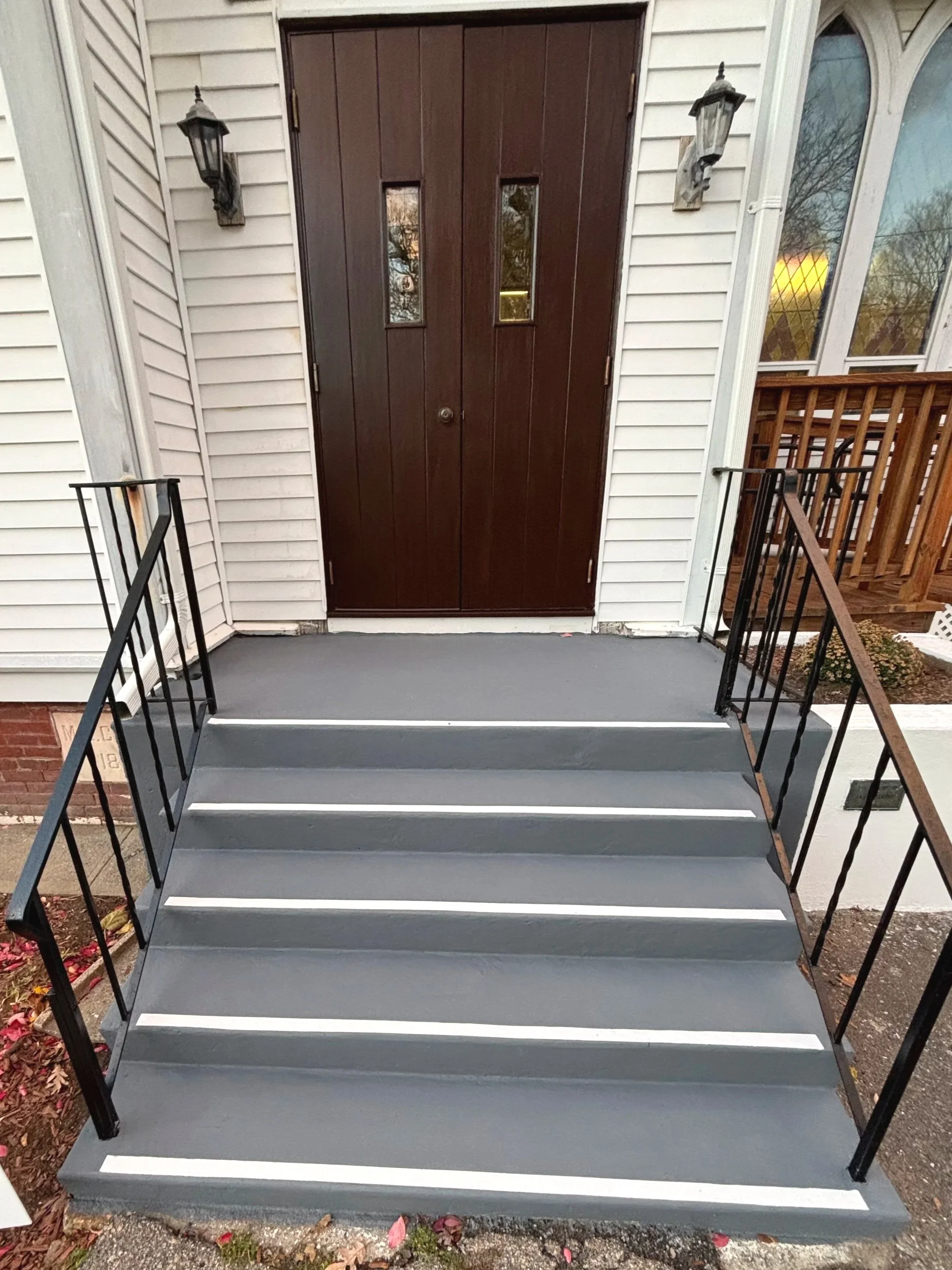 Front entrance steps with black metal railings leading to a dark wooden front door with two small windows, outside wall with white siding, two wall-mounted outdoor lanterns, and a nearby porch with a wooden railing.