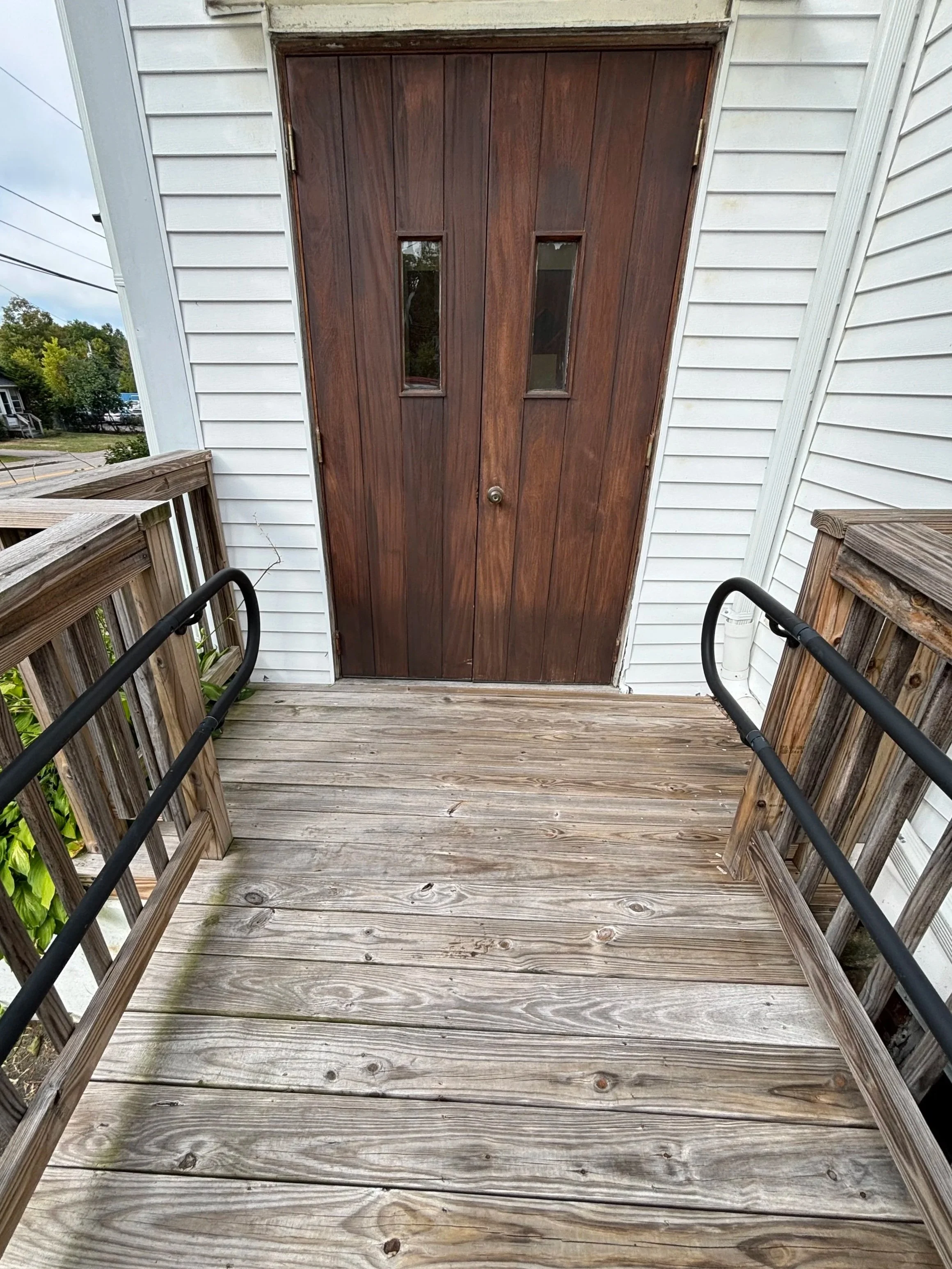 View of a wooden porch with railings leading up to a brown double door with two small vertical windows, attached to a white house.