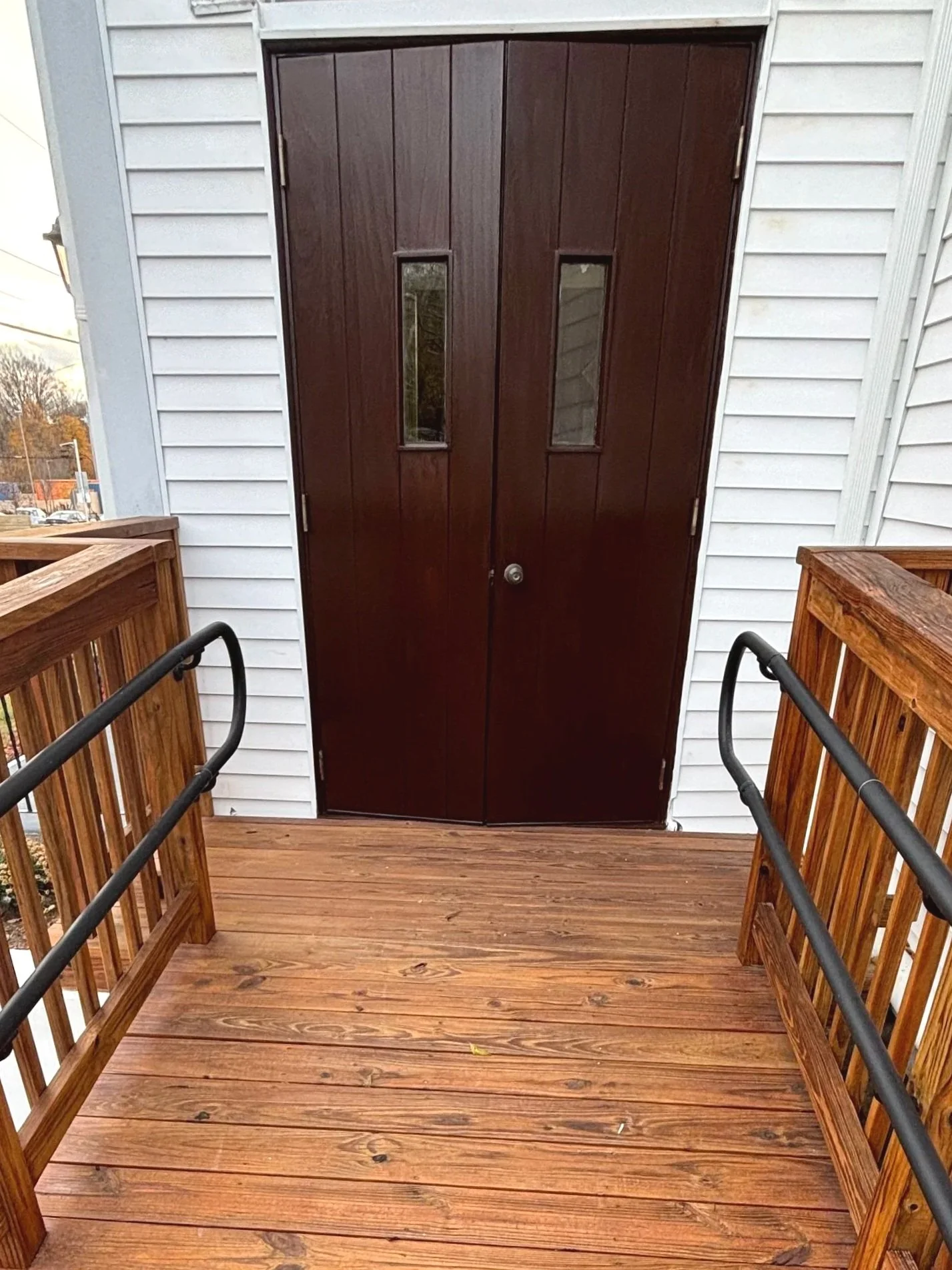 Wooden porch leading to a dark brown double door with small vertical windows, attached to a white house with horizontal siding.