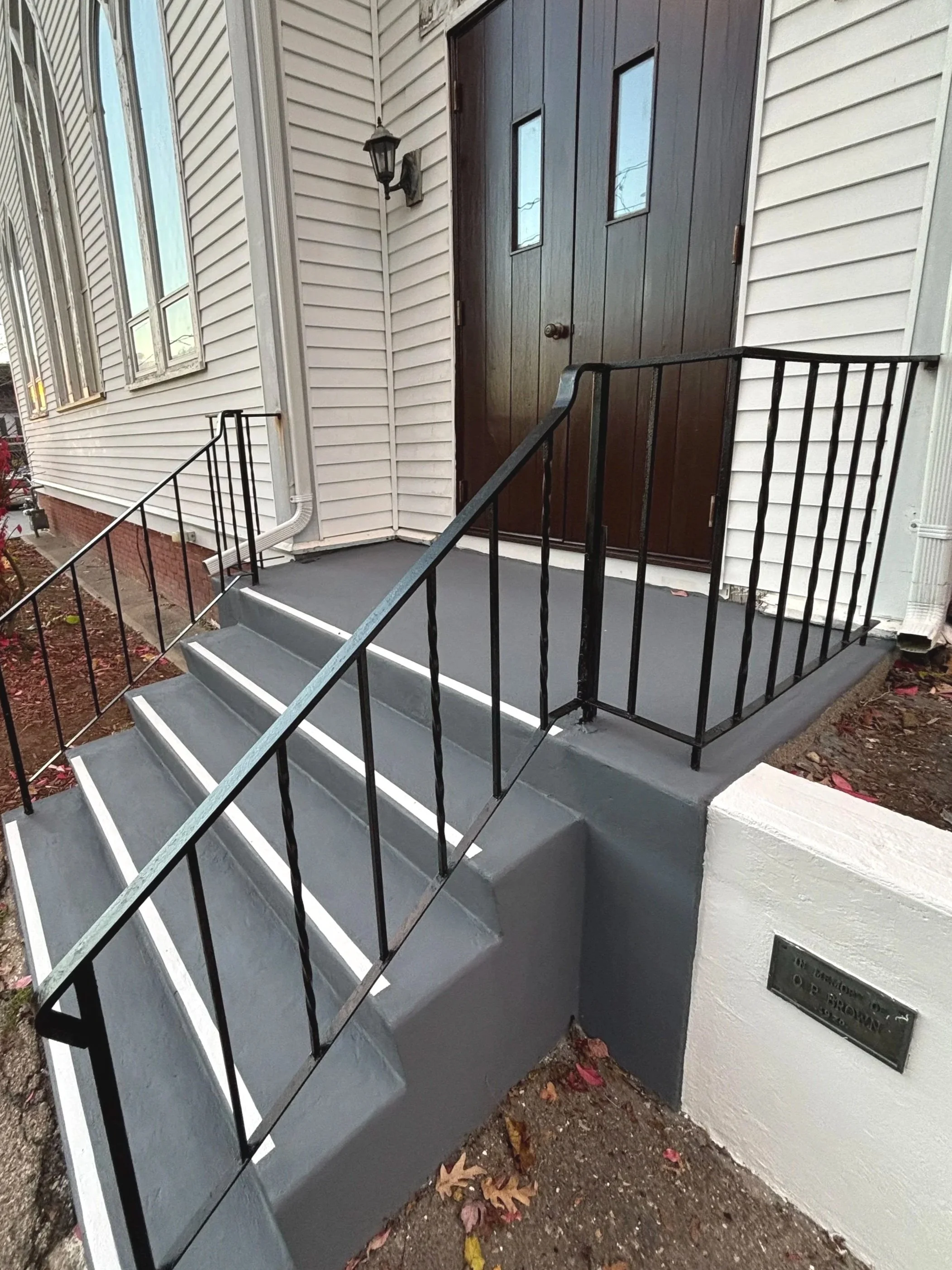 Entrance with stairs, black metal handrails, grey concrete porch, brown wooden door, white vinyl siding, outdoor light fixture, and a brass plaque on the white wall.