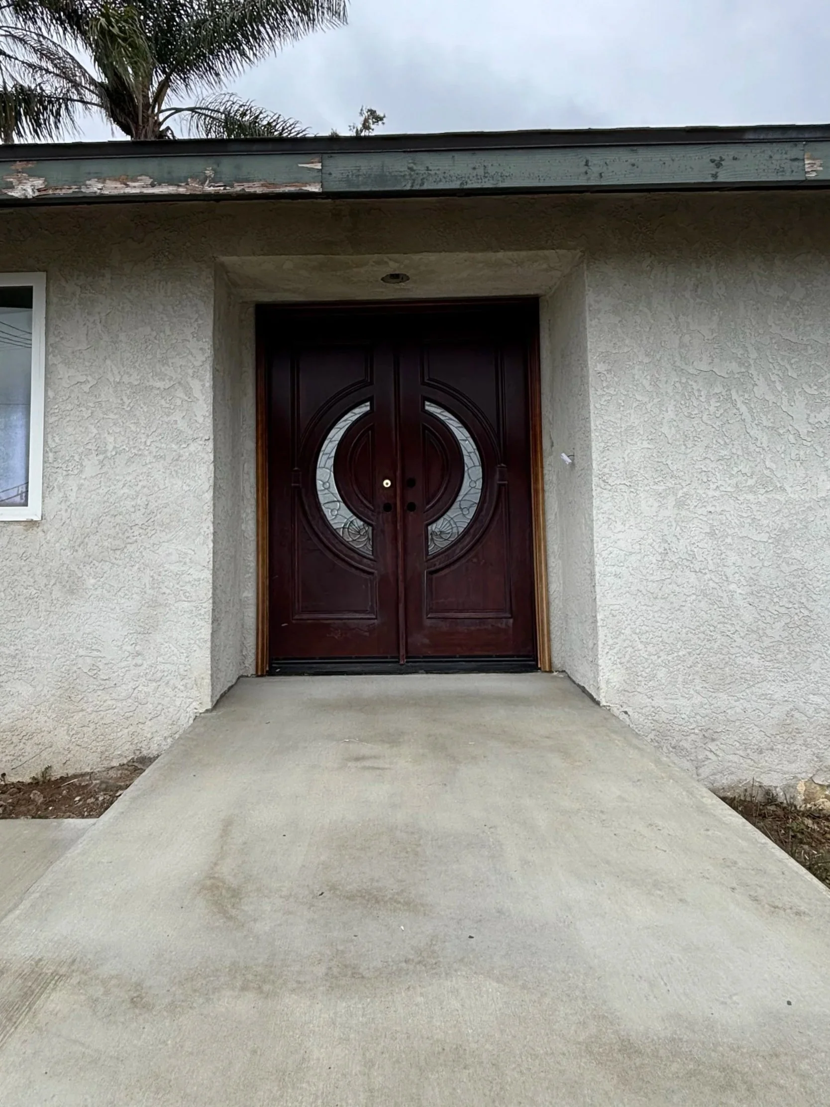 Front view of a house with a dark wooden double door and a small step ramp leading up to it, surrounded by stucco walls and a window on the left, with overcast sky and palm trees in the background.