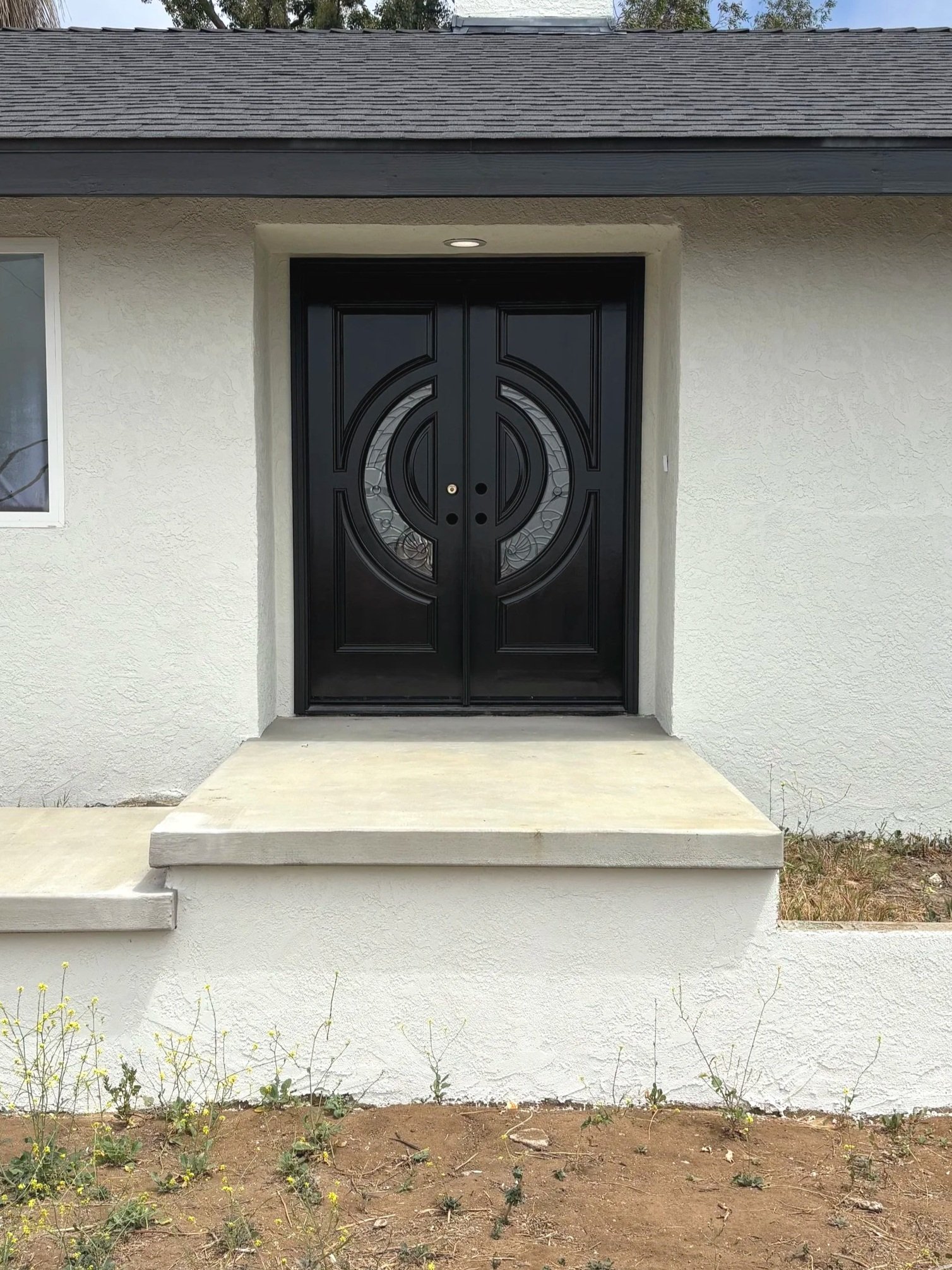 Black double door with decorative glass panels leading to the entrance of a house, surrounded by white stucco walls and a small concrete porch.