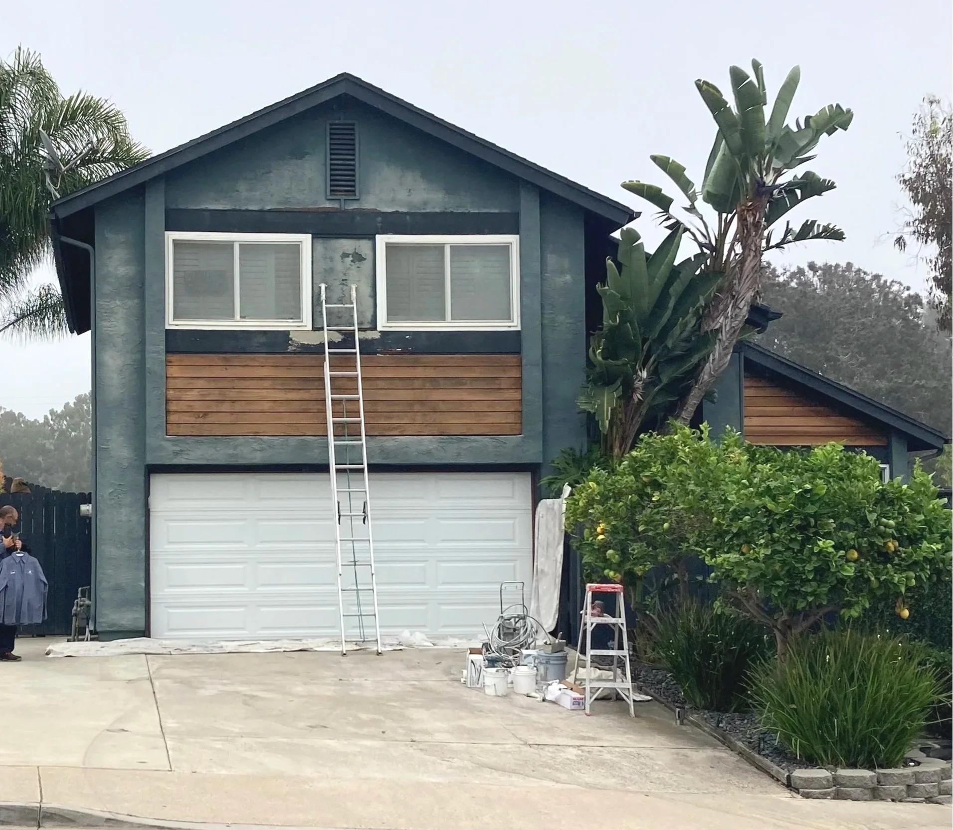 Front view of a two-story house under renovation with a ladder leaning against the second floor, construction tools and supplies on the driveway, and a person on the left holding a shirt.